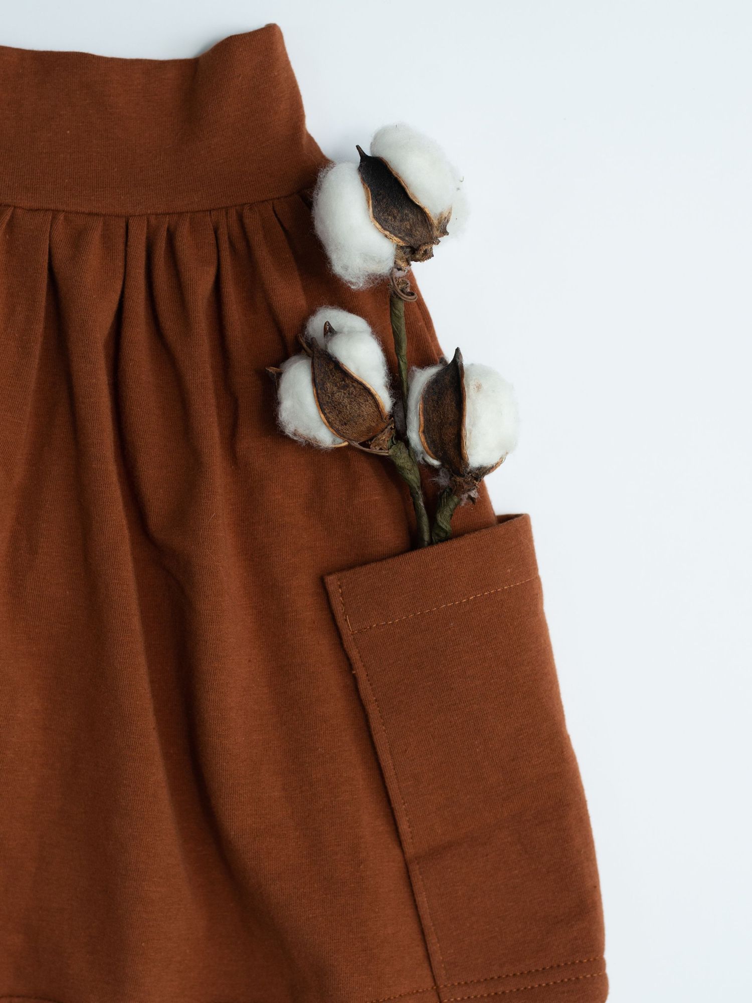 Brown fabric garment with pleated waistline detail and pocket, decorated with natural cotton bolls against white background.