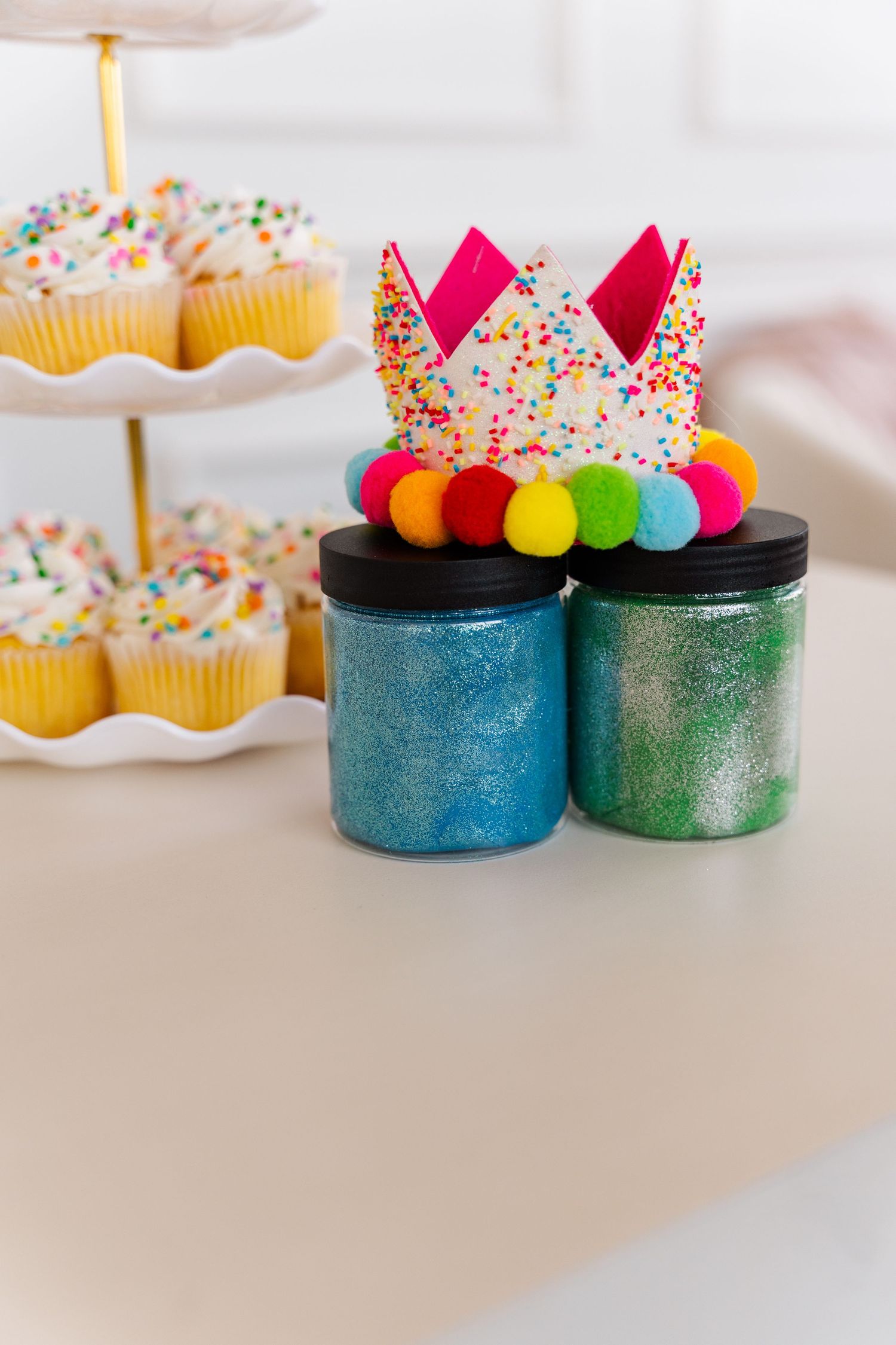 Colorful cupcake decorations with sprinkles and rainbow candy balls displayed on sparkly blue and green containers.