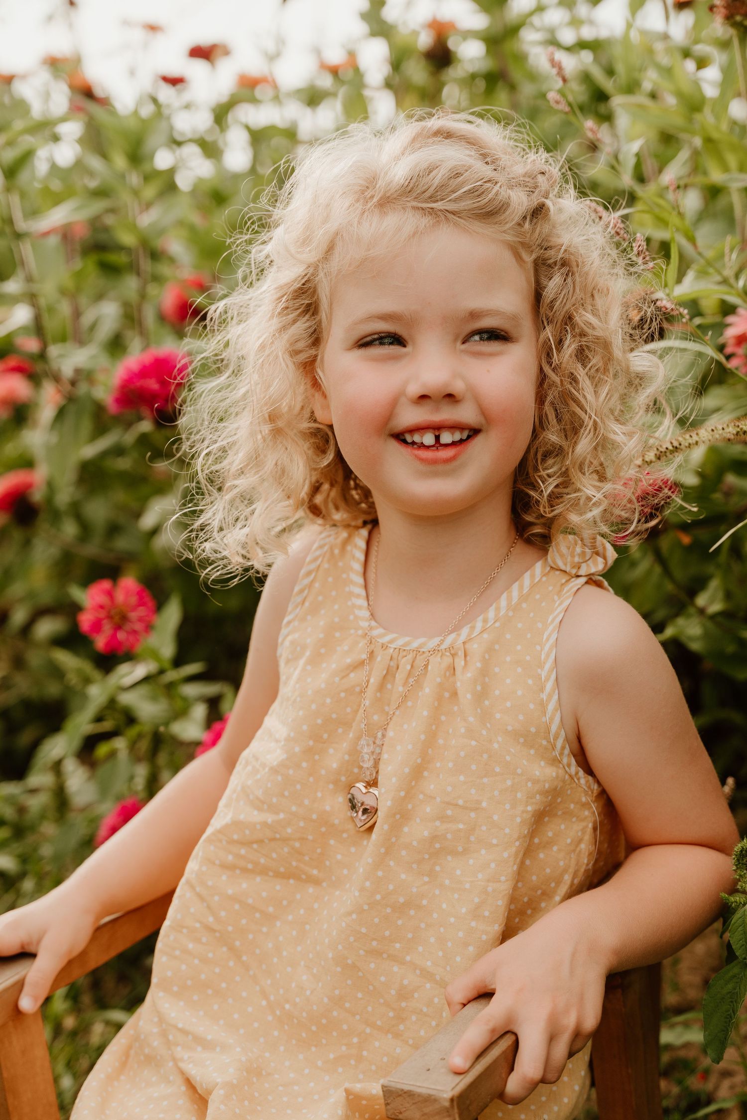 Smiling young girl with curly blonde hair wearing a yellow dress, sitting outdoors among red flowers.
