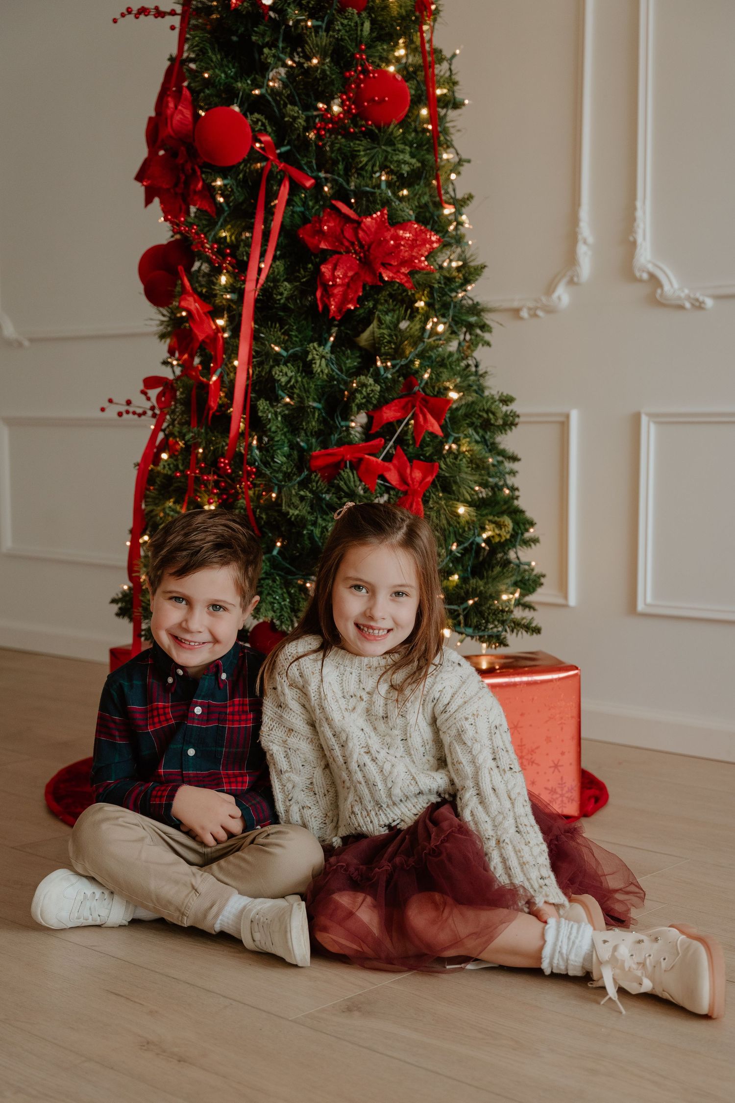 Two children sit together in front of a decorated Christmas tree with red ornaments and bows on hardwood flooring.