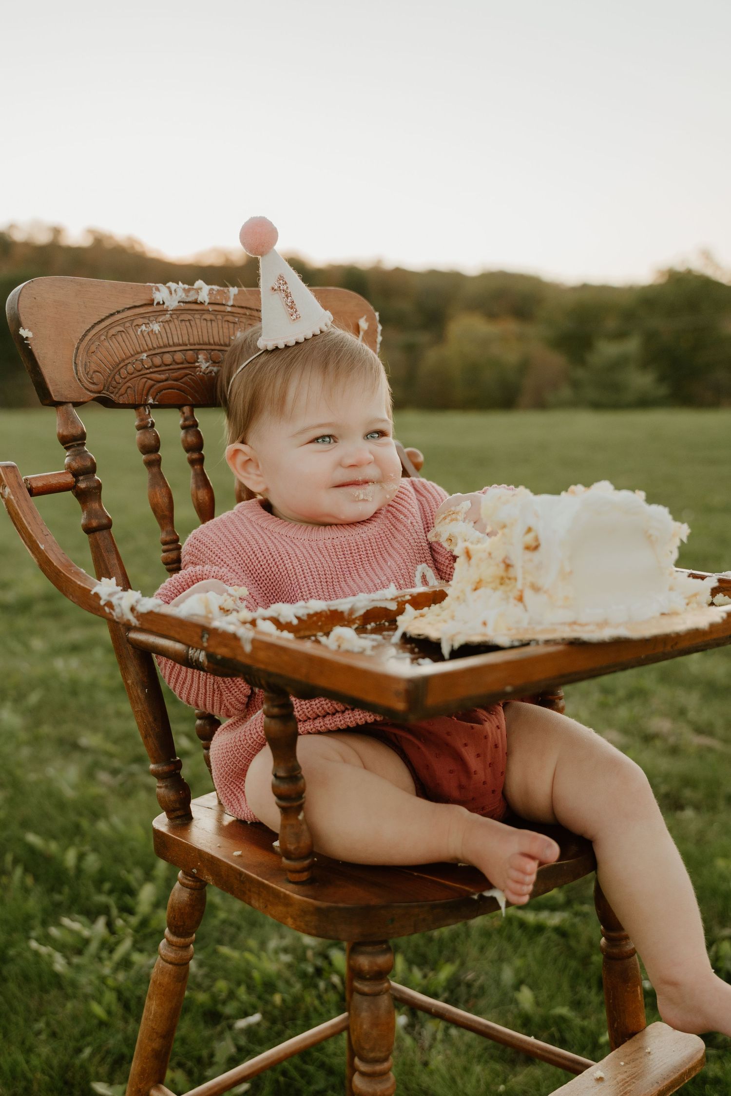 Baby in pink sweater sits in wooden antique chair with birthday cake smash setup during golden hour outdoor photo session.