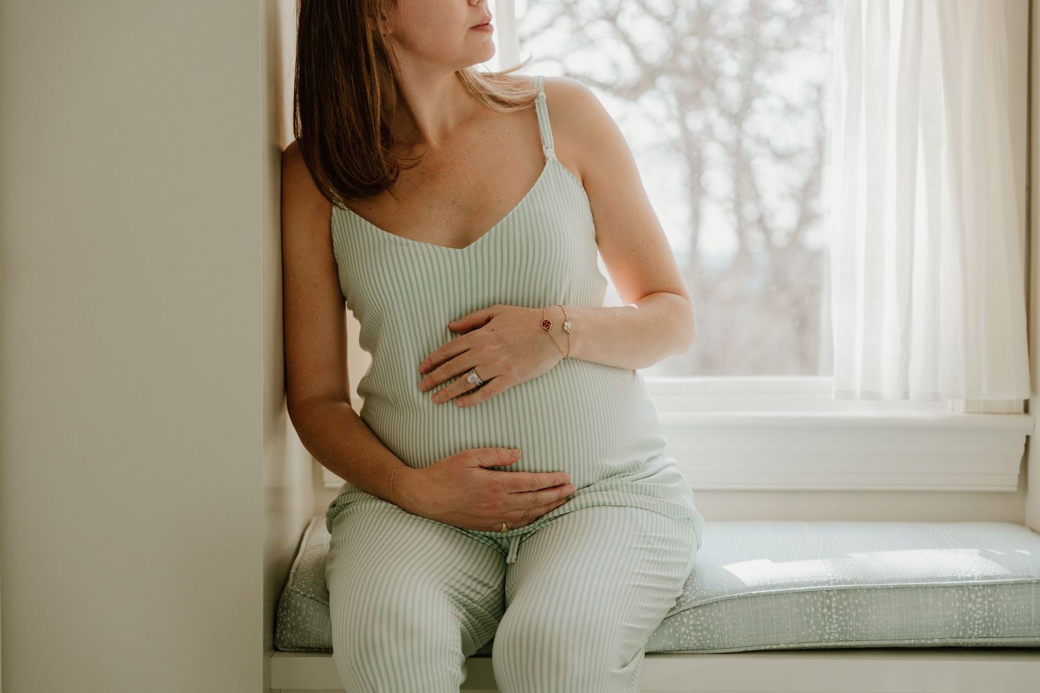 A woman in mint green loungewear sits by a window while cradling her pregnant belly.