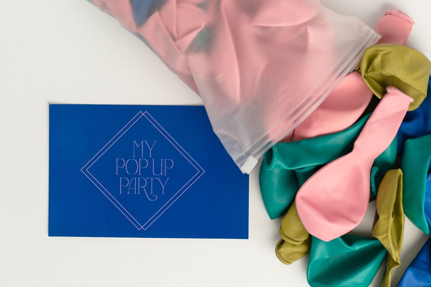A blue party invitation card with diamond logo lies beside pink, green and gold latex balloons.