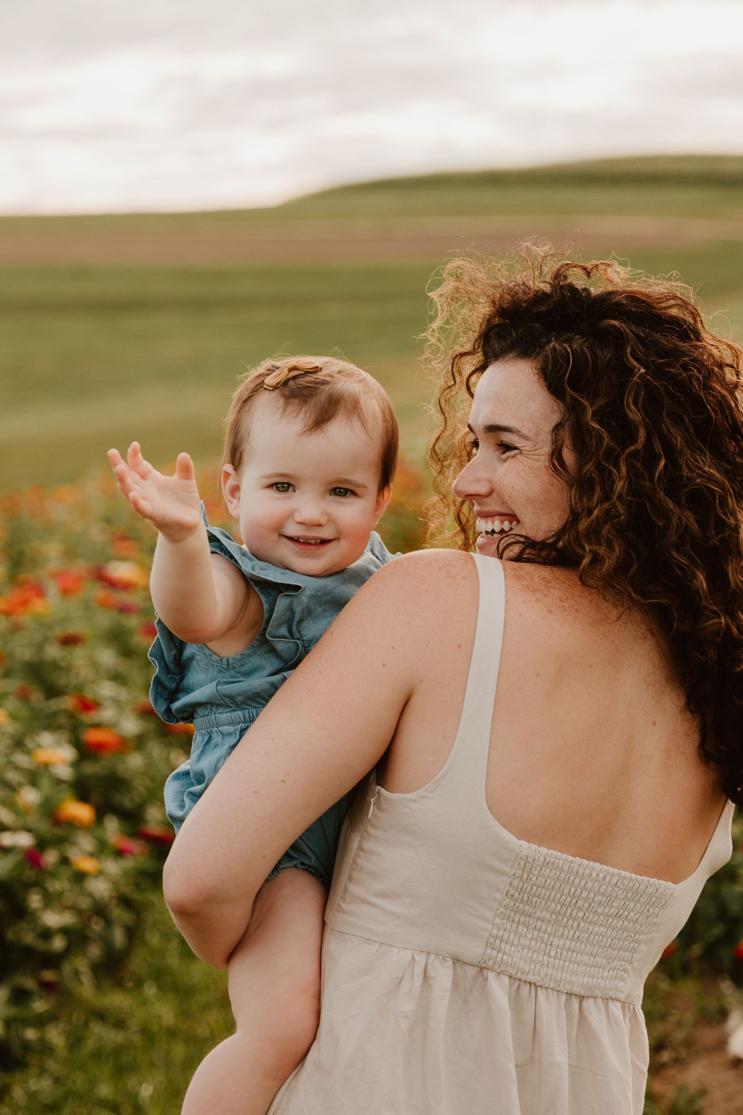 A curly-haired woman in a white dress holds a smiling baby in a denim outfit in a colorful flower field.