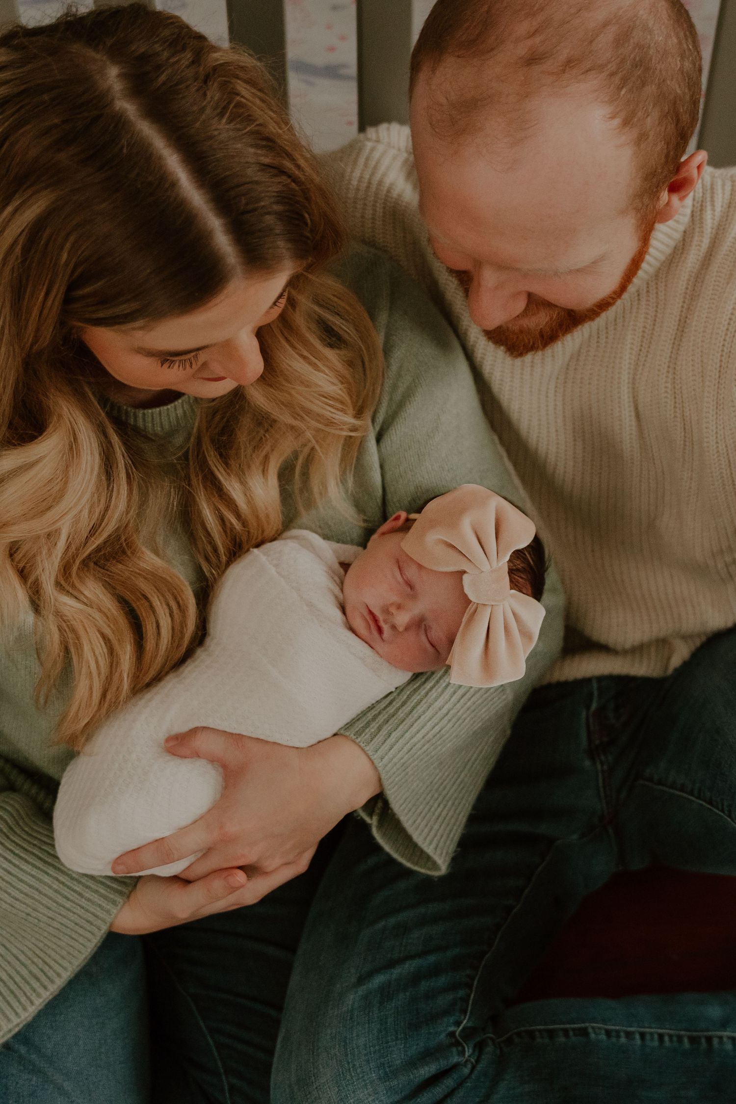 A sleeping newborn baby wearing a white bow headband is cradled lovingly by parents in matching sage green sweaters.