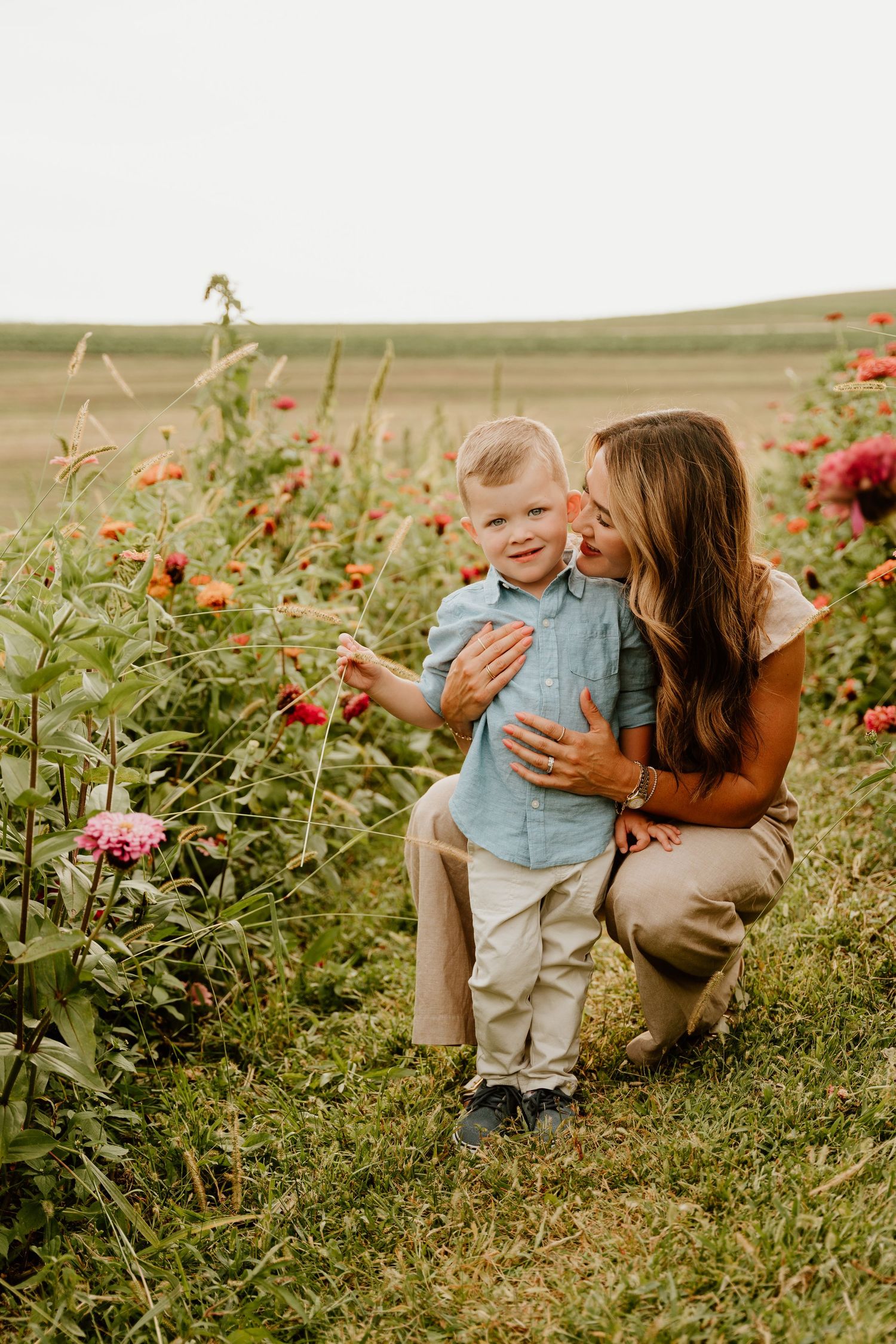 A mother crouches beside her toddler son in a vibrant flower field, sharing a tender moment among pink and red blooms.