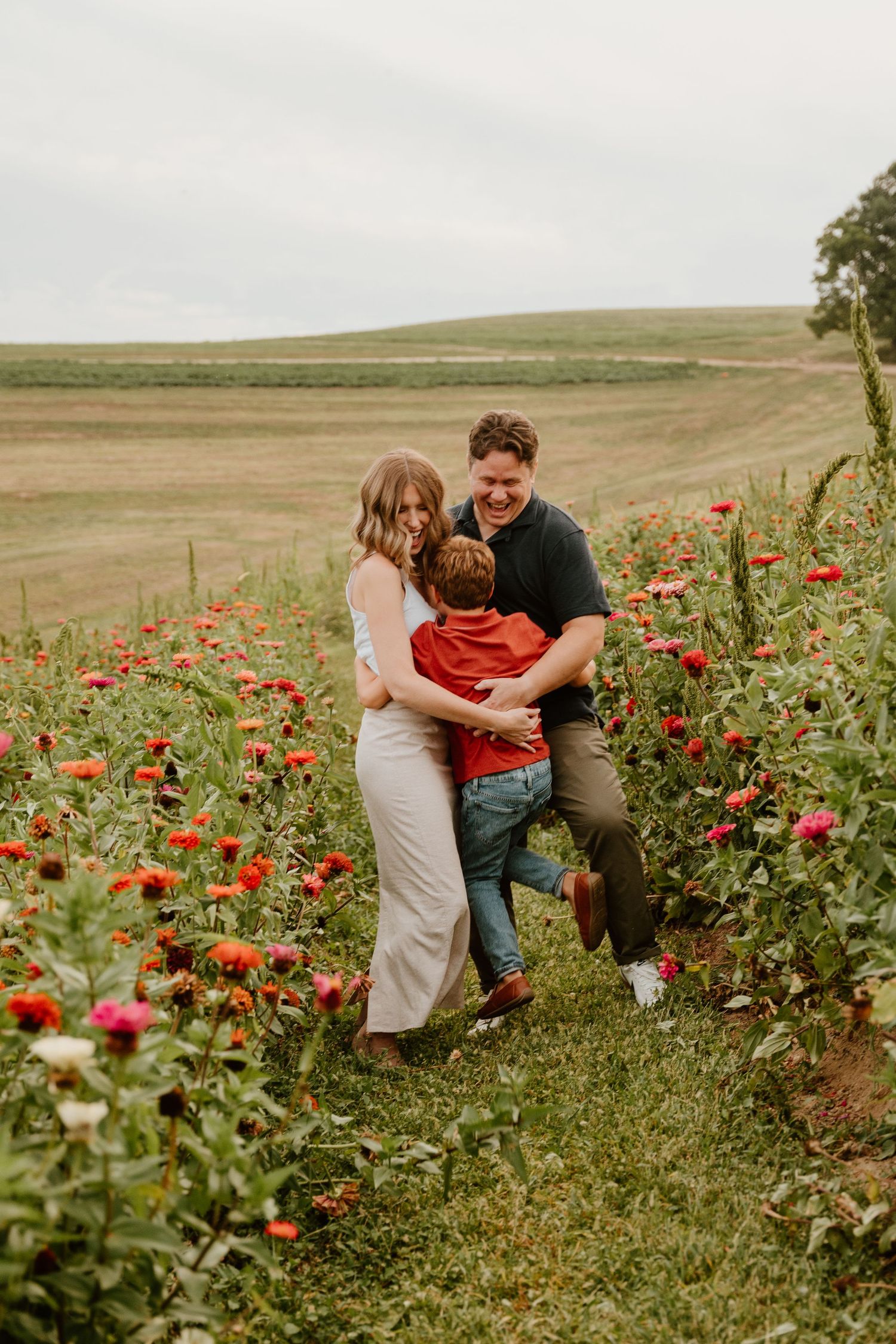 Family of three laughing together in a vibrant flower field with red and pink blooms on a cloudy day.