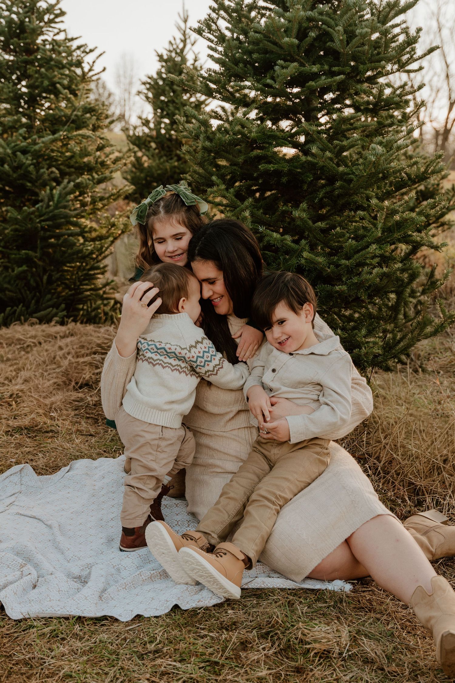 A family in neutral colored clothing sits together on a white blanket near evergreen trees in an outdoor setting.