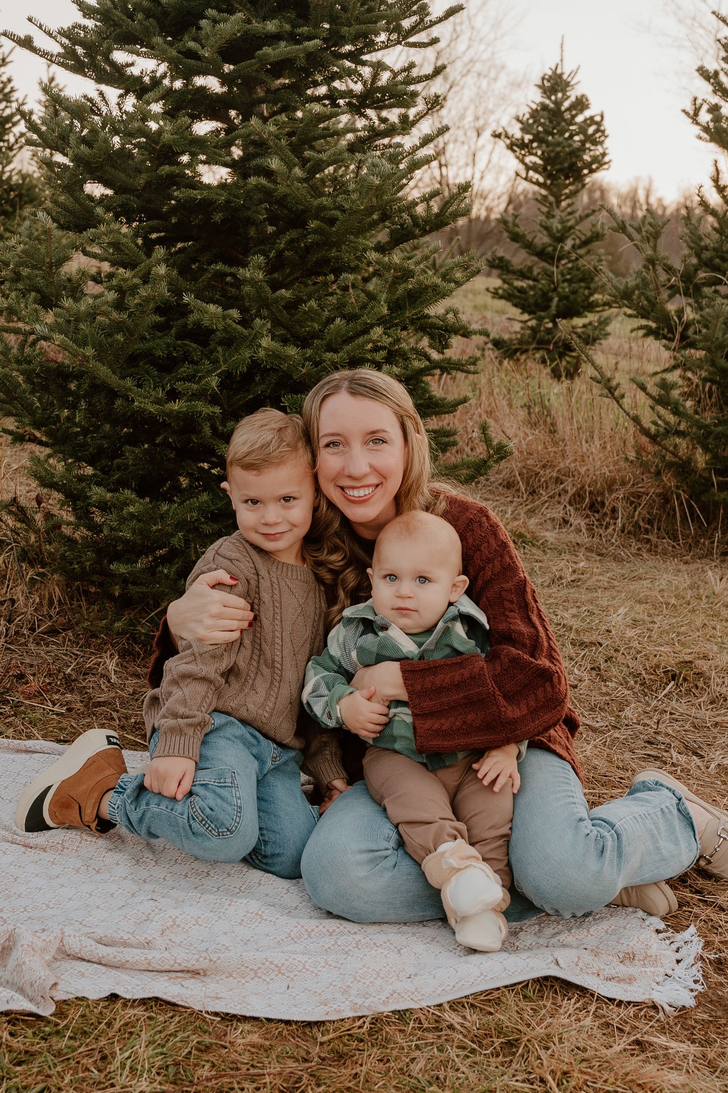 A family sits together on a white blanket outdoors among evergreen trees in warm earth-toned clothing.