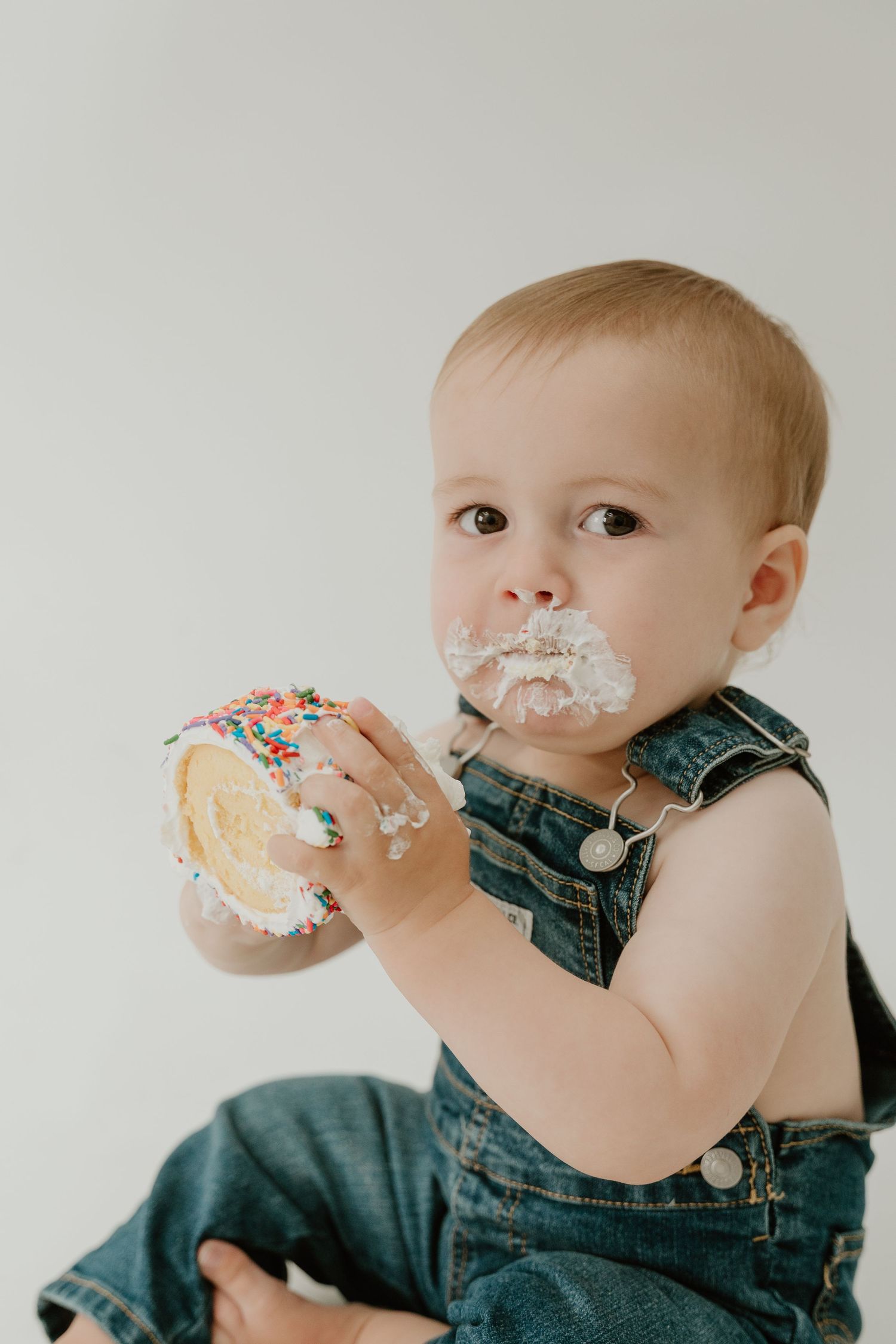 Baby in denim overalls enjoying a messy first taste of birthday cake in a series of candid shots.