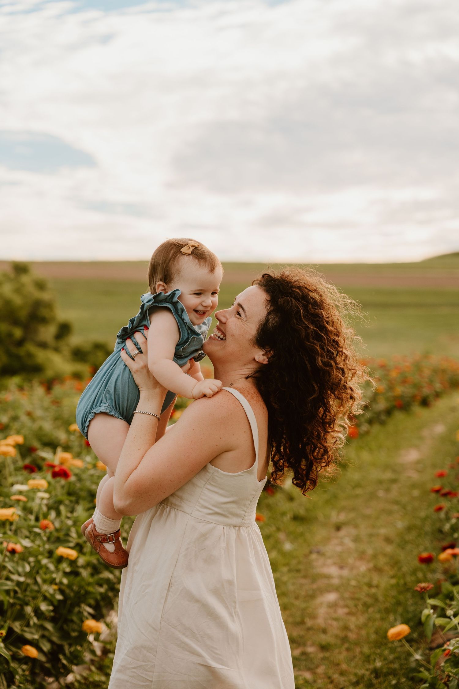 Joyful mother lifting smiling baby girl in a colorful zinnia flower field under cloudy skies.