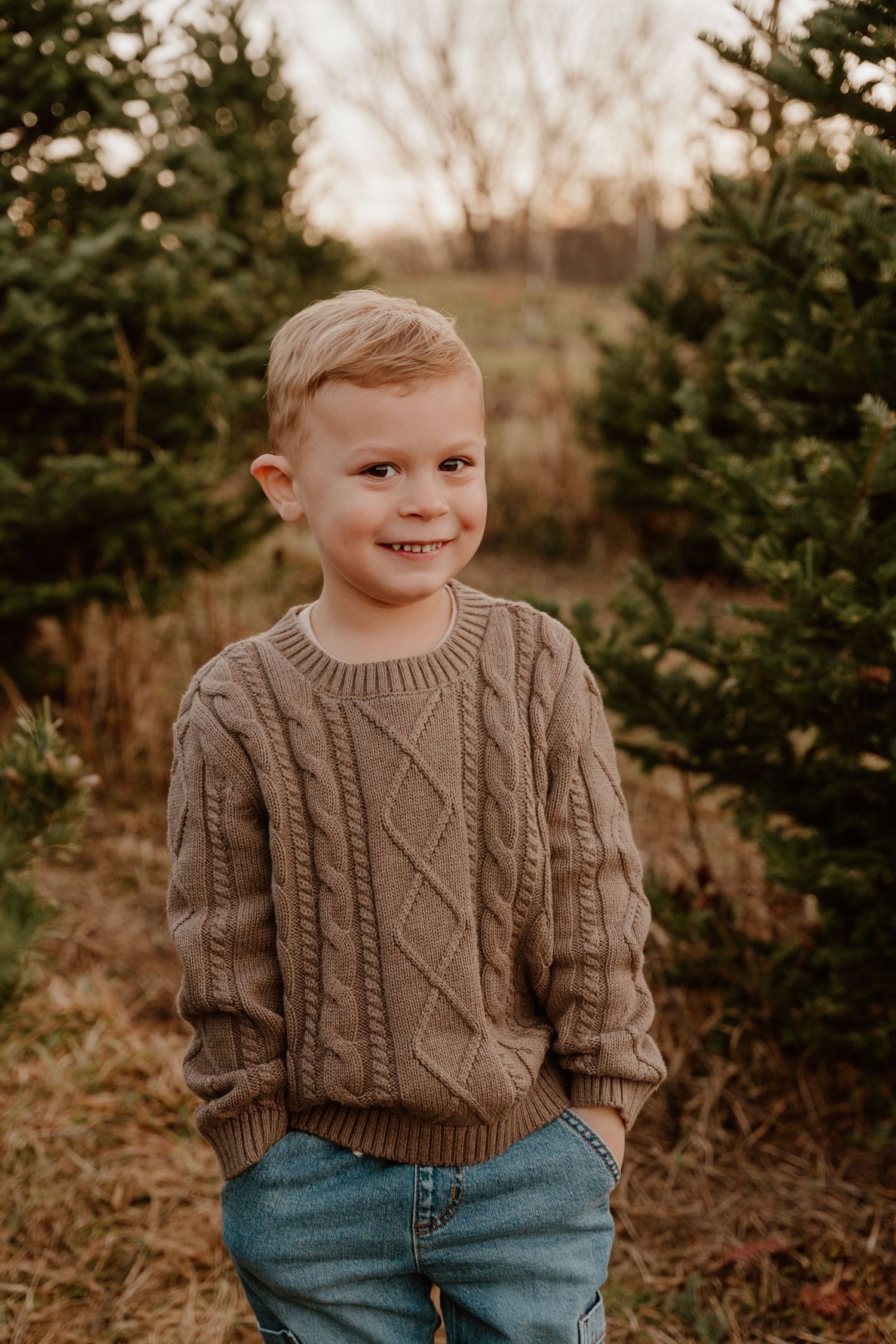 Child wearing brown cable knit sweater and jeans smiles while standing among evergreen trees at sunset.