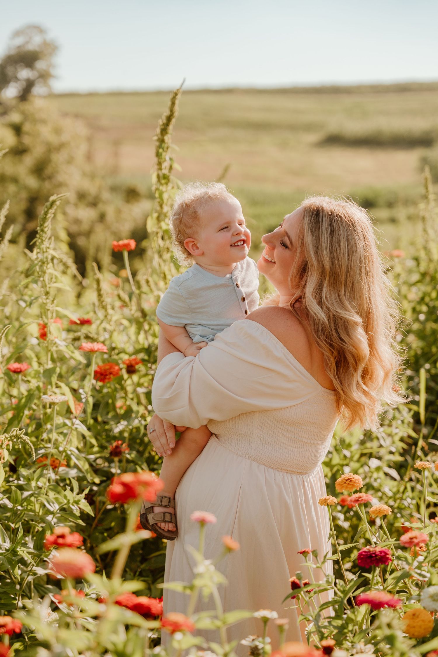 A smiling mother in a white dress holds her laughing baby boy in a vibrant field of red and orange flowers.