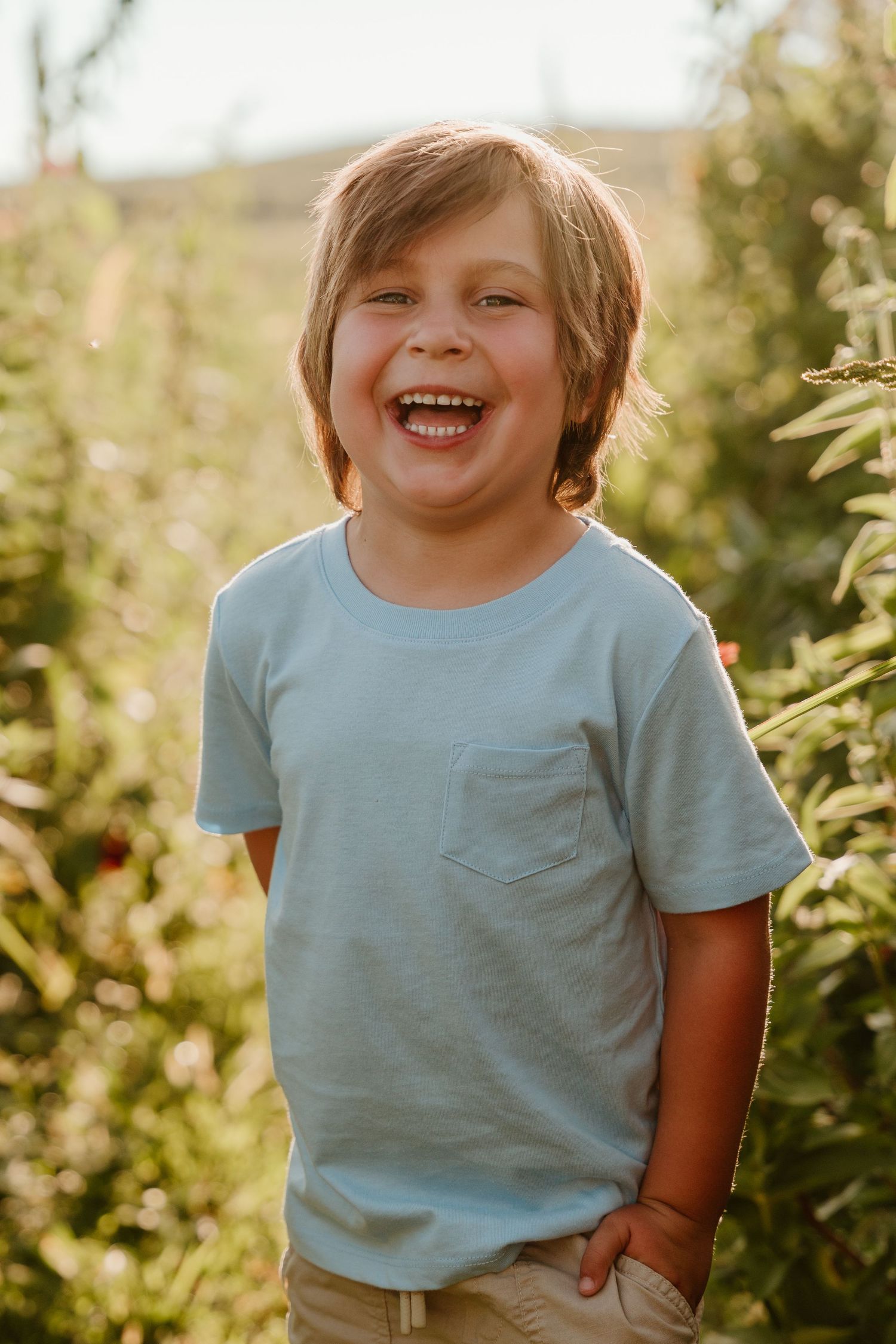 A laughing young boy in a light blue t-shirt stands outdoors in a sunny apple orchard.