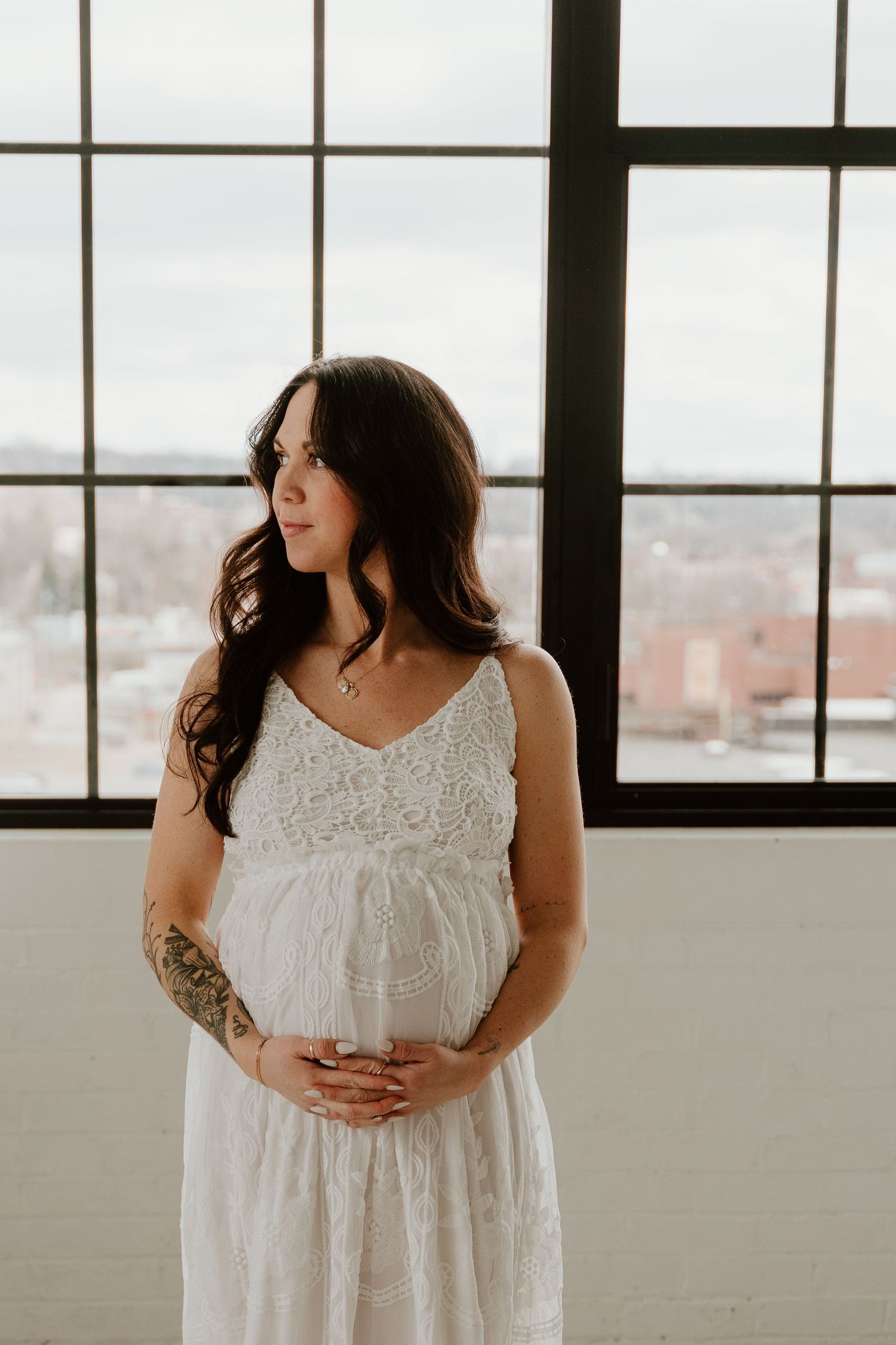 Expectant woman in white lace dress stands by large industrial window with city view while holding her baby bump.