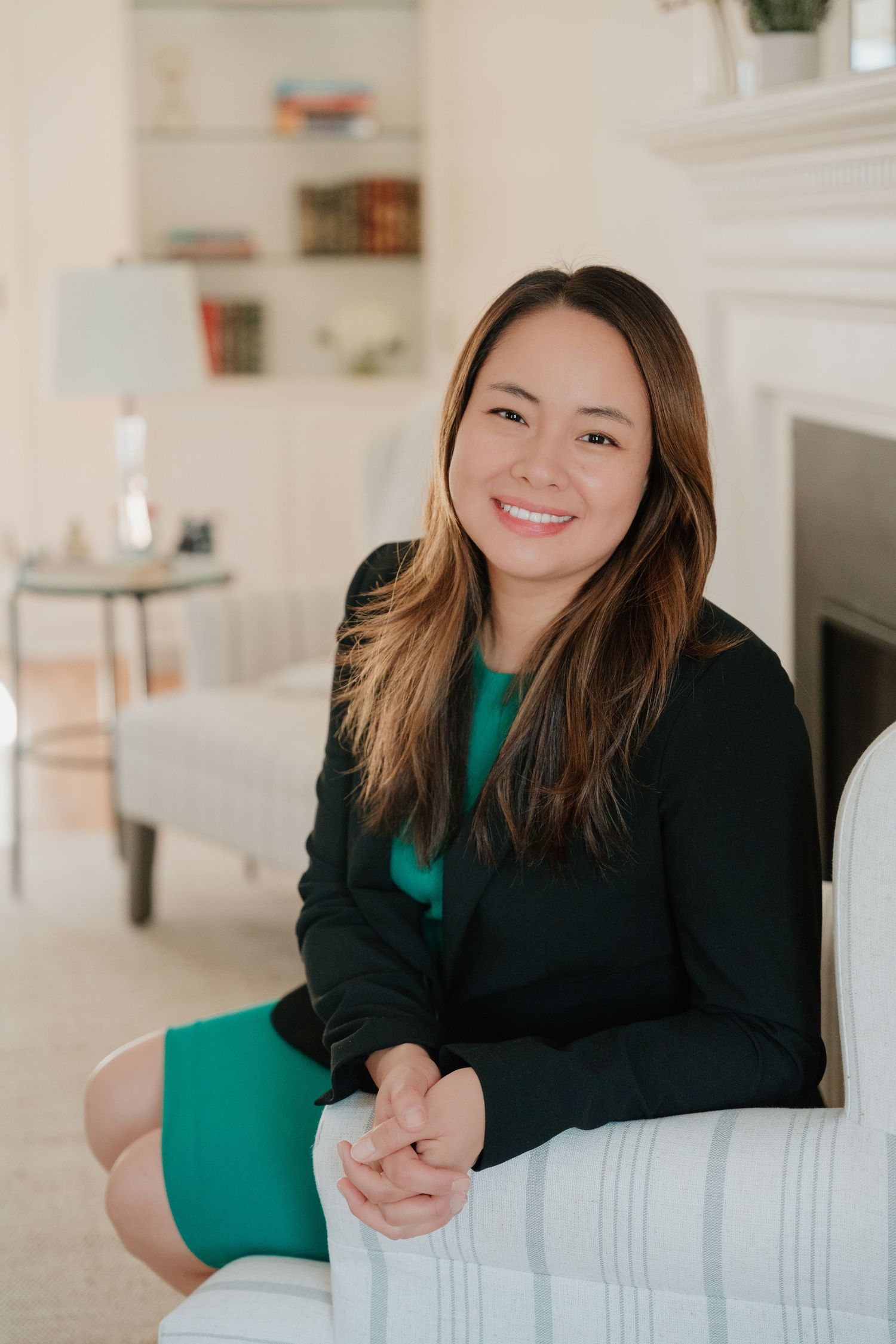 Professional wearing black blazer and emerald green dress poses casually against white interior wall.