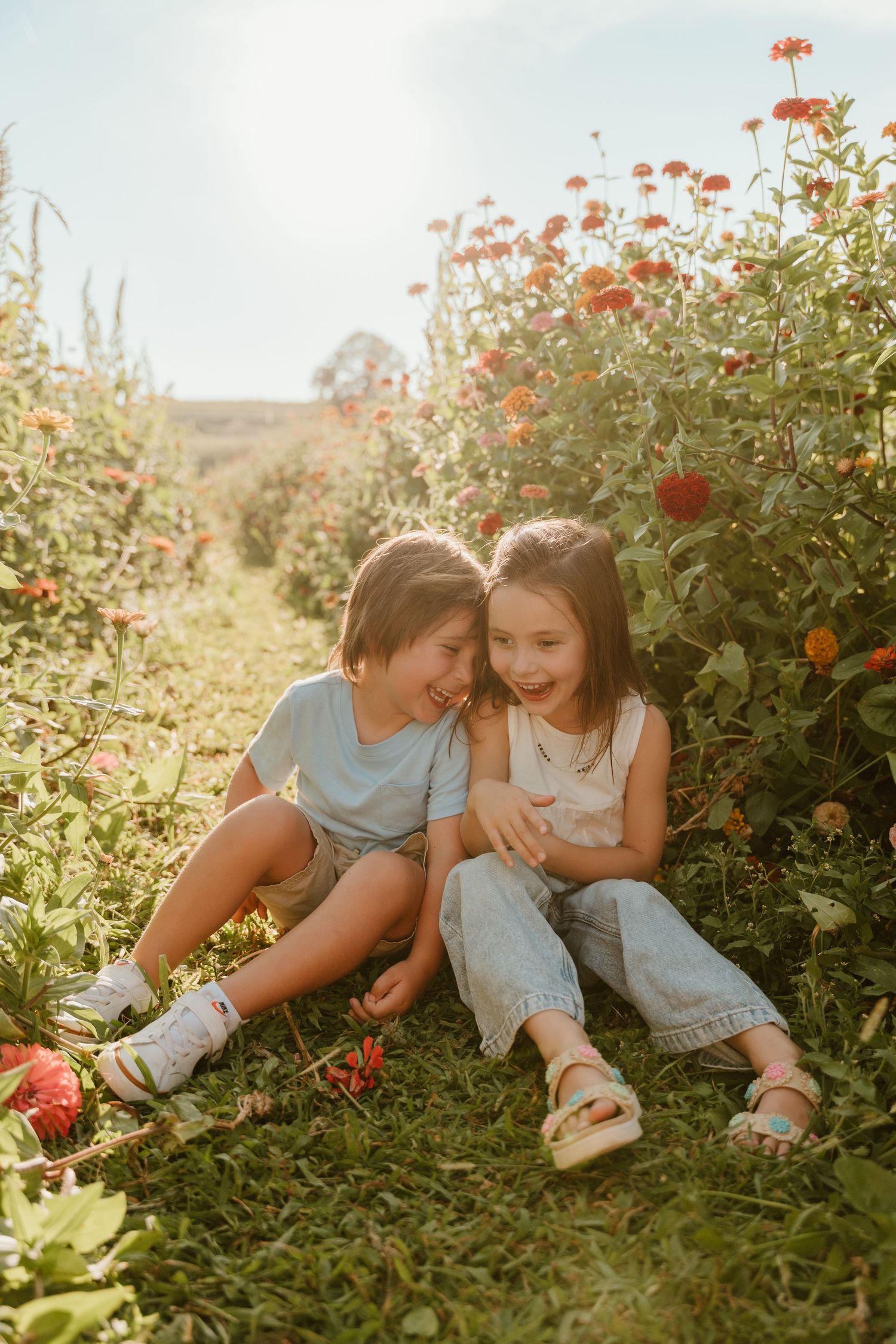 Two children sit together smiling in a sunny flower field with red blooms, wearing casual summer outfits.