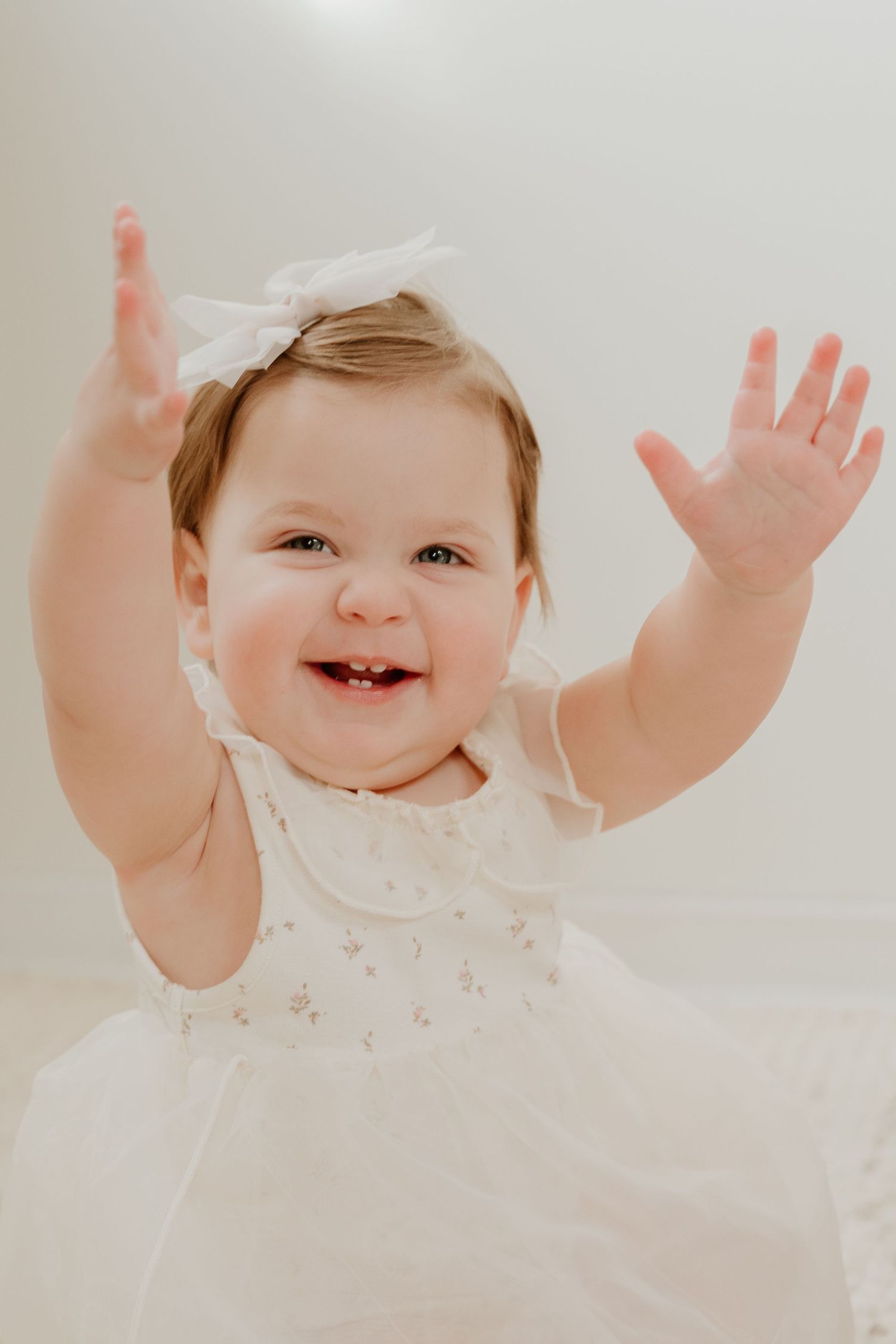A joyful baby in a white dress raises their arms up while laughing against a light background.