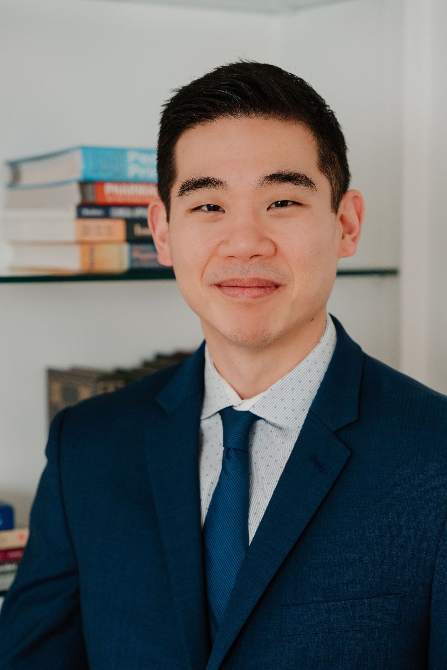 Professional portrait of someone in a navy suit and patterned tie smiling warmly against a light background with books.