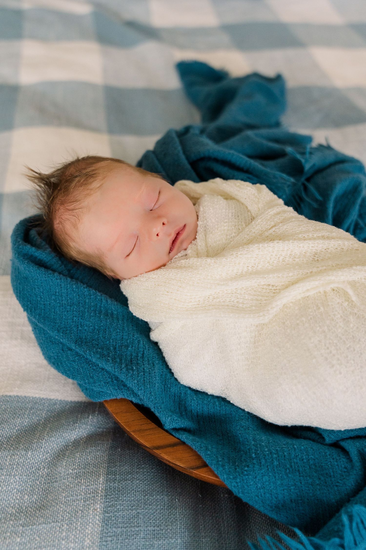 Sleeping newborn wrapped in white swaddle on blue plaid bedding.