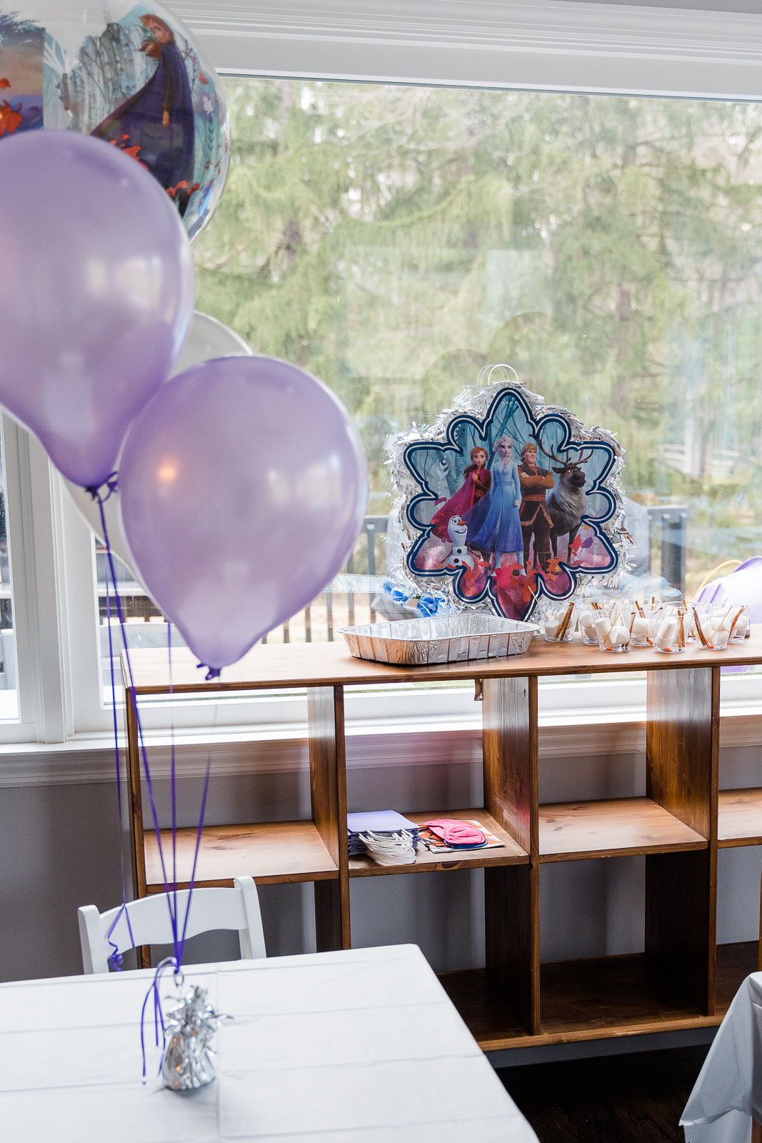 Frozen-themed party setup with purple balloons and decorative display shelves near a window.