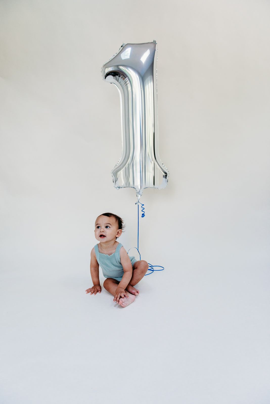 Baby in turquoise outfit sitting with silver number 1 balloon against white background.
