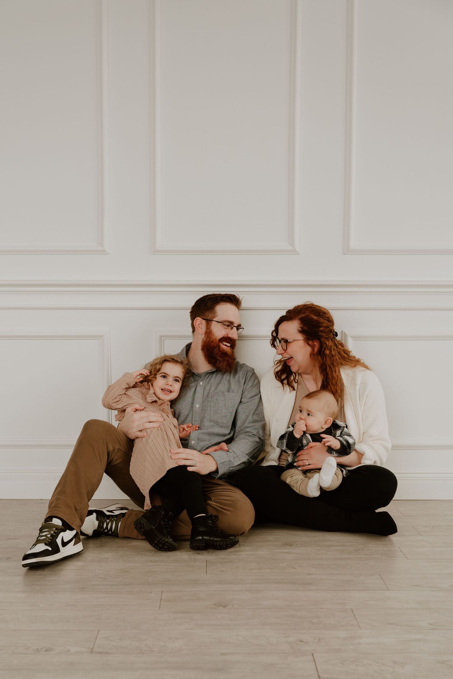 Family sitting together on wooden floor against white wall wearing casual clothing in soft natural light.