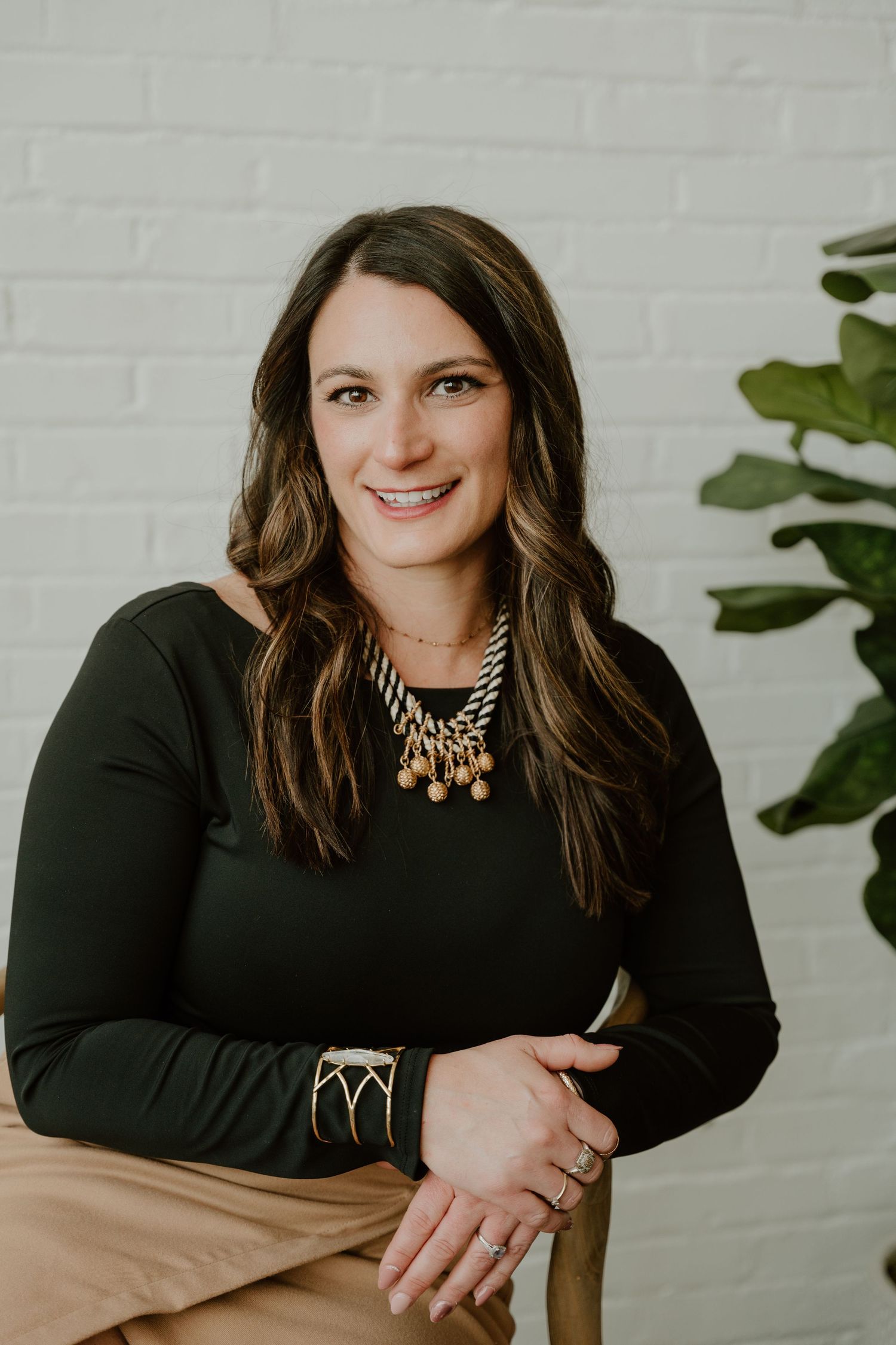 Professional portrait of a person in a black long sleeve top and gold statement necklace against a white brick wall.