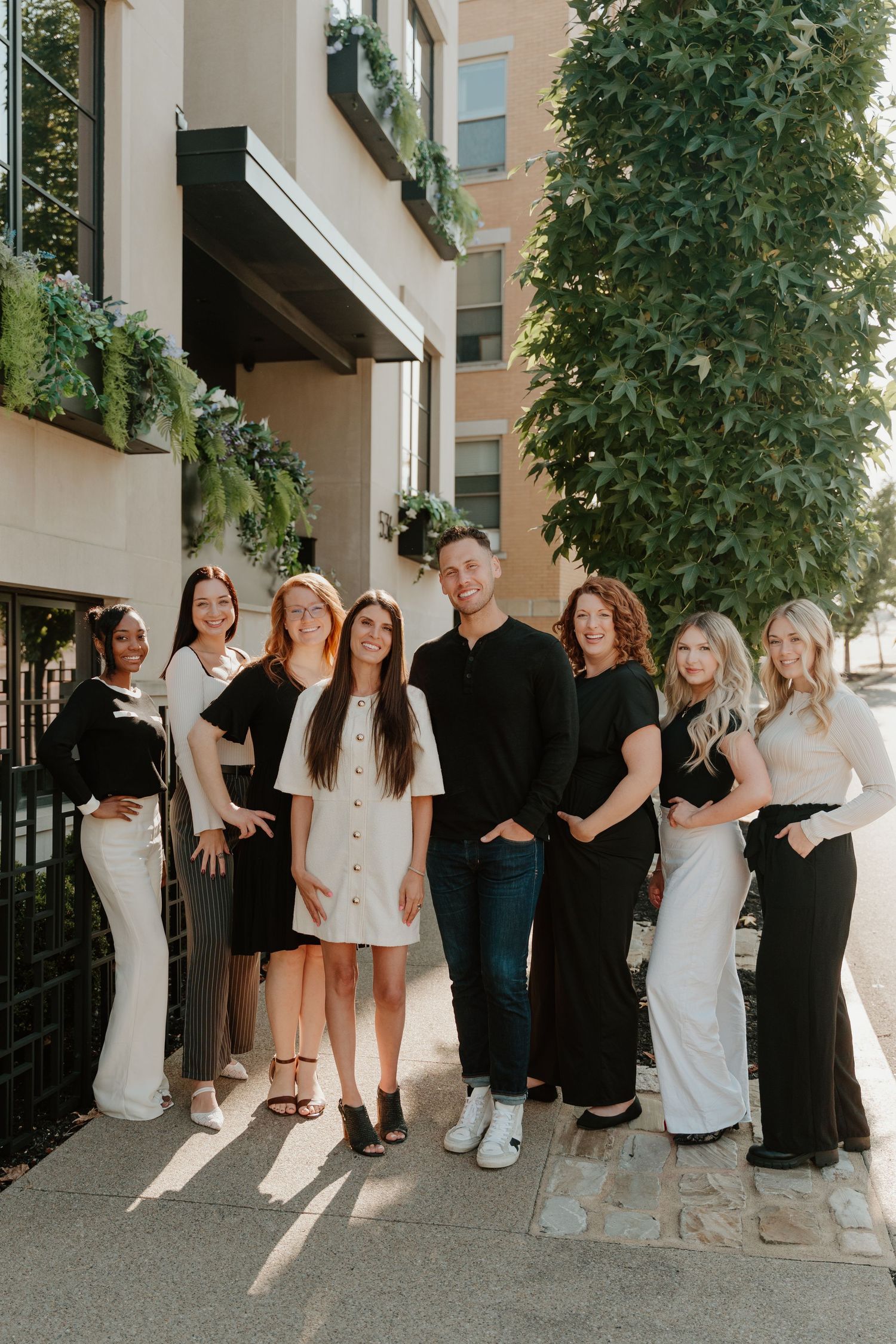 A group of people in black and white outfits pose together in a casual outdoor setting with greenery in the background.