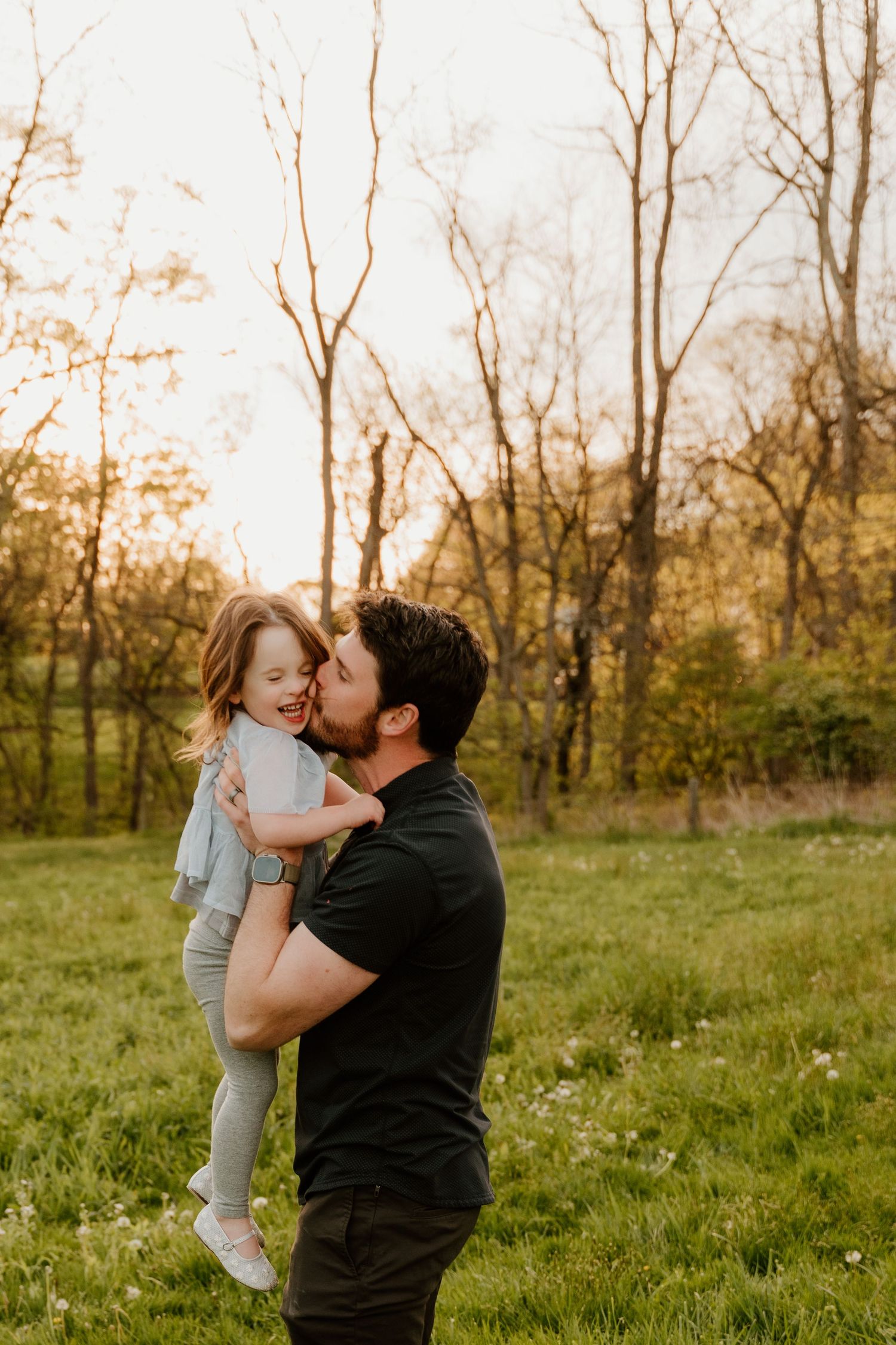 Sweet family moment captured in green field during golden hour with bare trees behind.