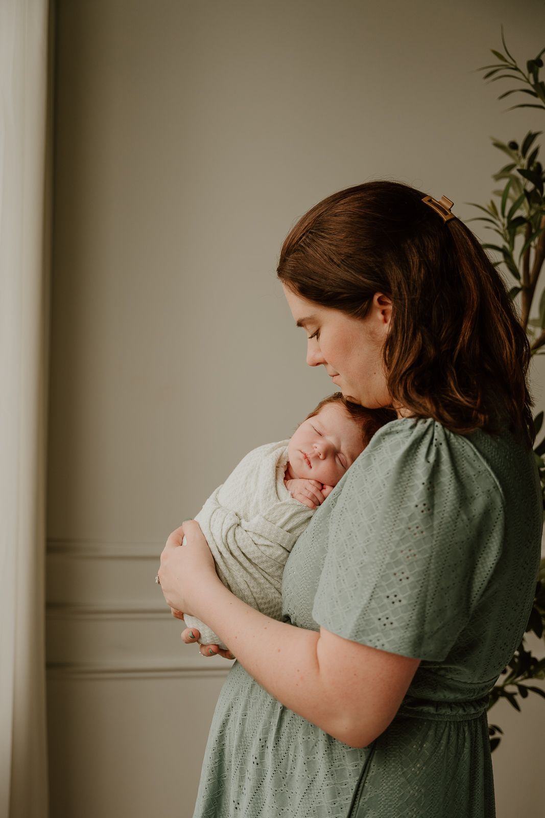 A tender moment captured as a woman in a sage green dress cradles a swaddled newborn baby against her chest.