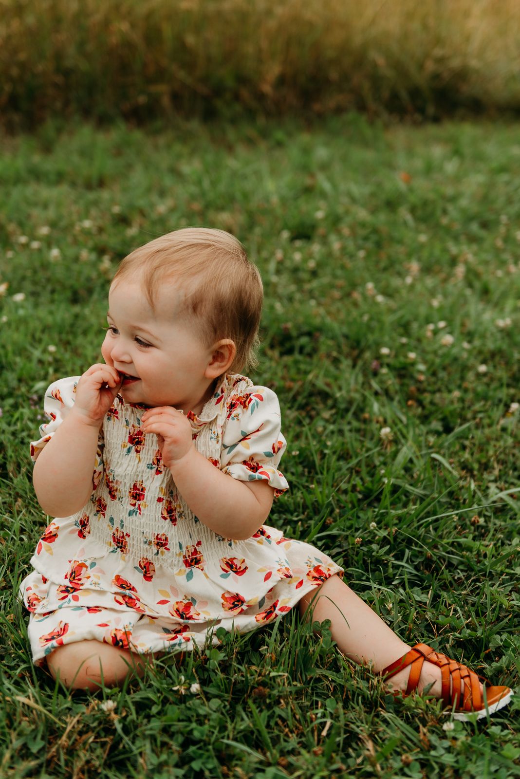 Child sitting in grass wearing floral dress and brown sandals.