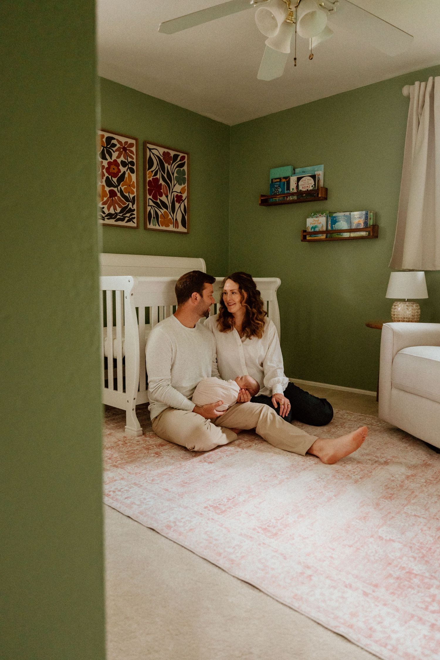 A couple sits together on a pink floral rug in a room with sage green walls and white furniture.