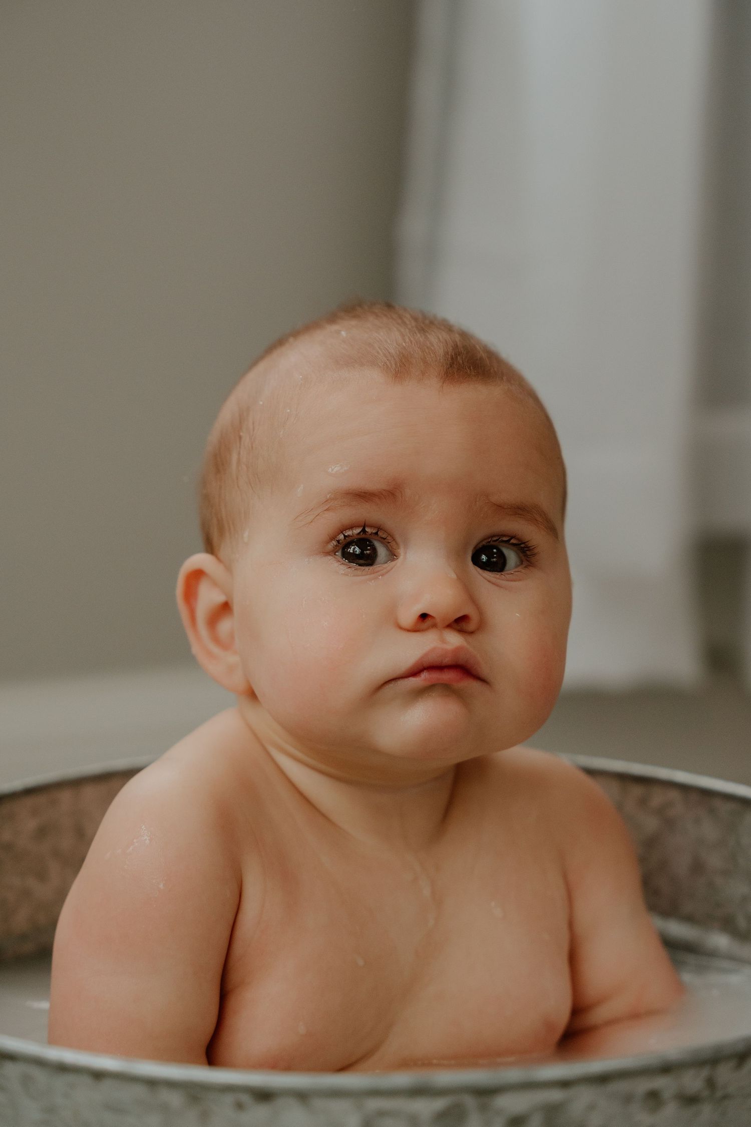 Sweet baby sits in vintage metal basin during bathtime with curious expression.