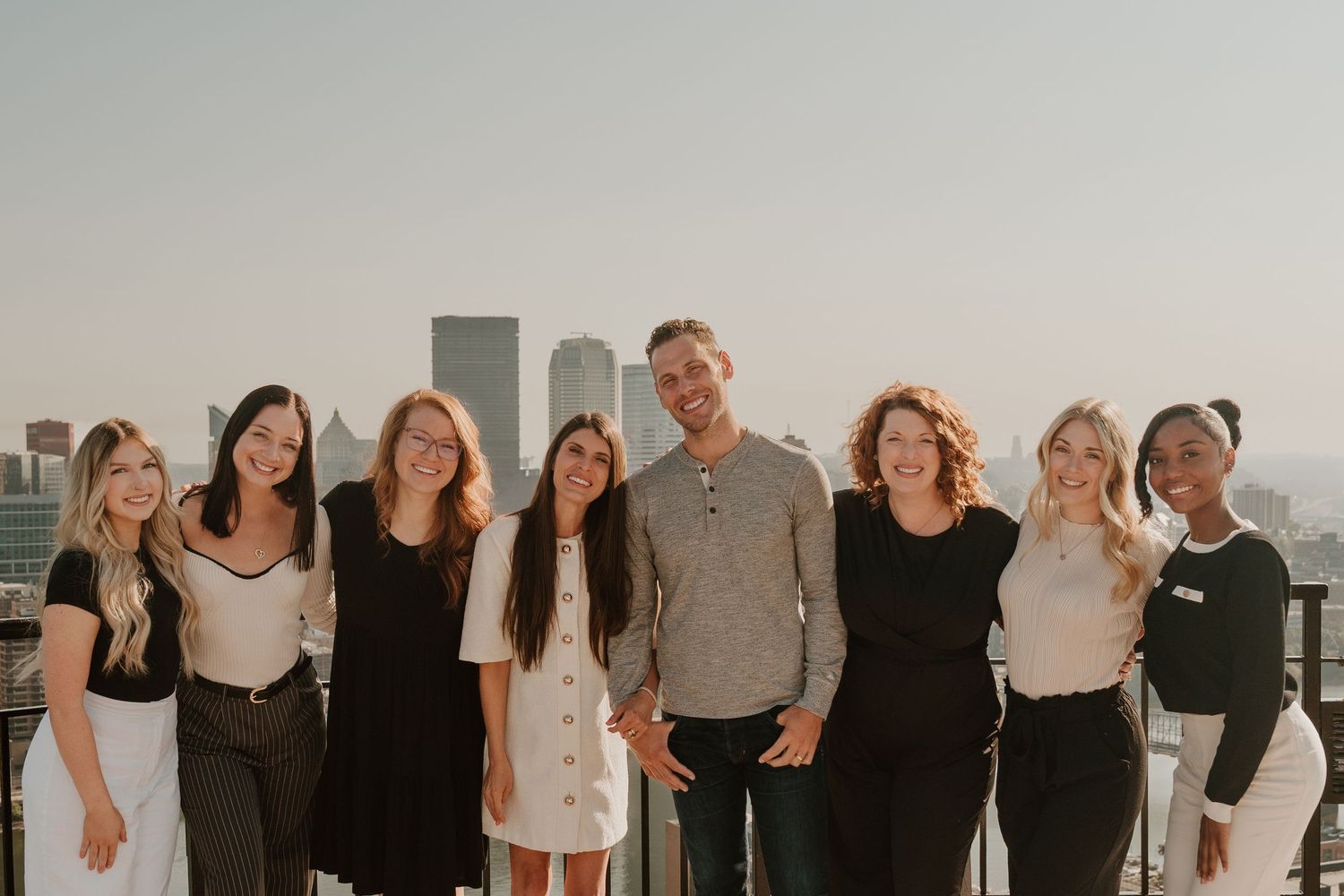 Group of people standing together in a row against a light colored wall posing for a casual photo.