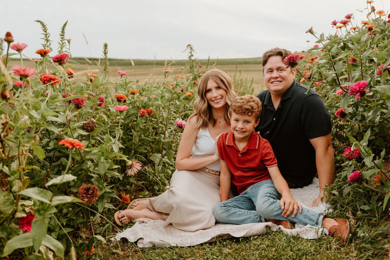 Family of three sitting in a vibrant flower field, smiling together on a white blanket surrounded by red blooms.