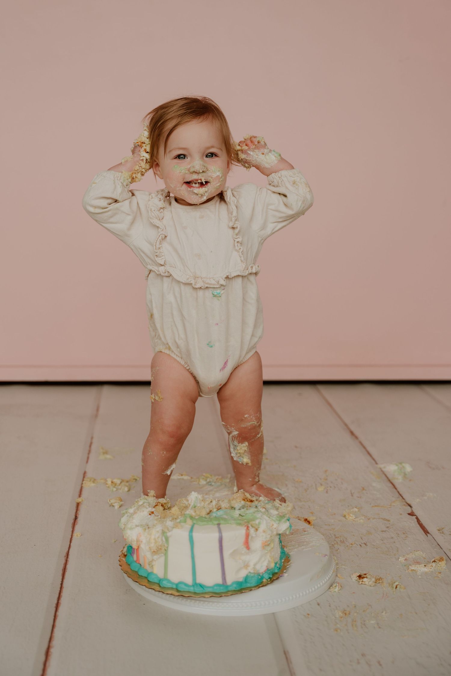A sequence of photos showing a baby in white clothing standing and playing with a smashed birthday cake on a light pink background.