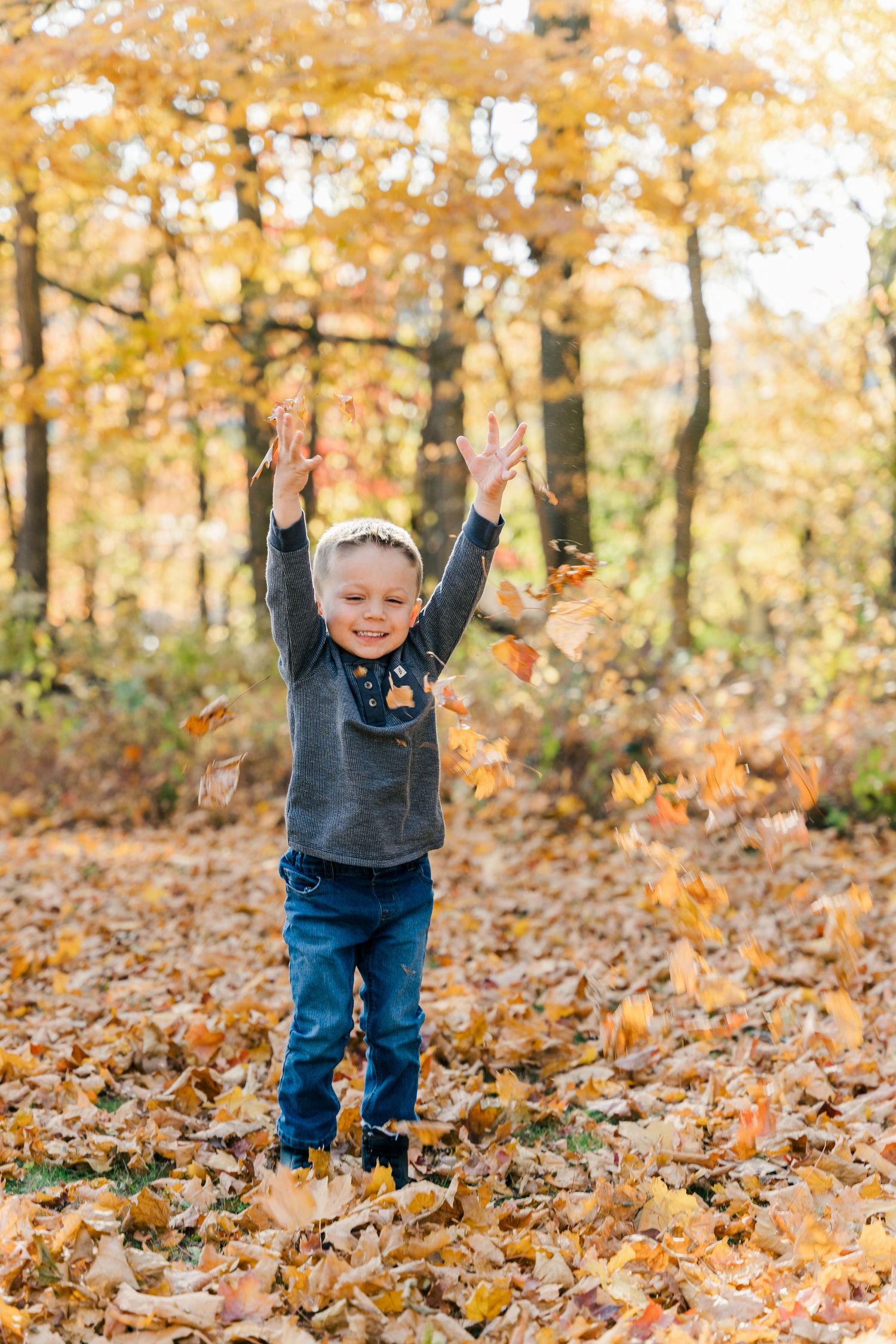 Child throwing autumn leaves in air during fall forest photoshoot.