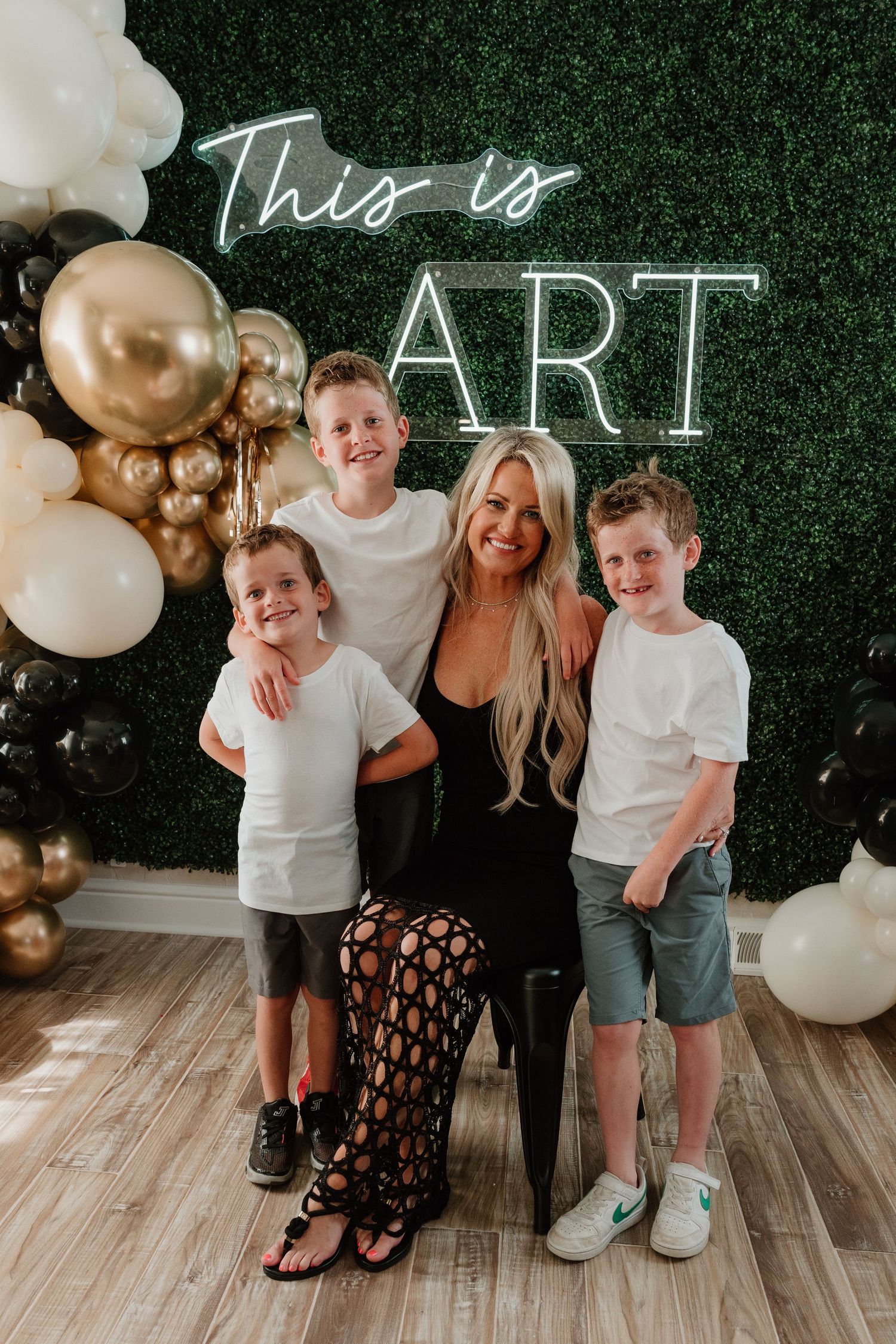 Family poses together against green hedge wall with white and gold balloon decorations at photo session.