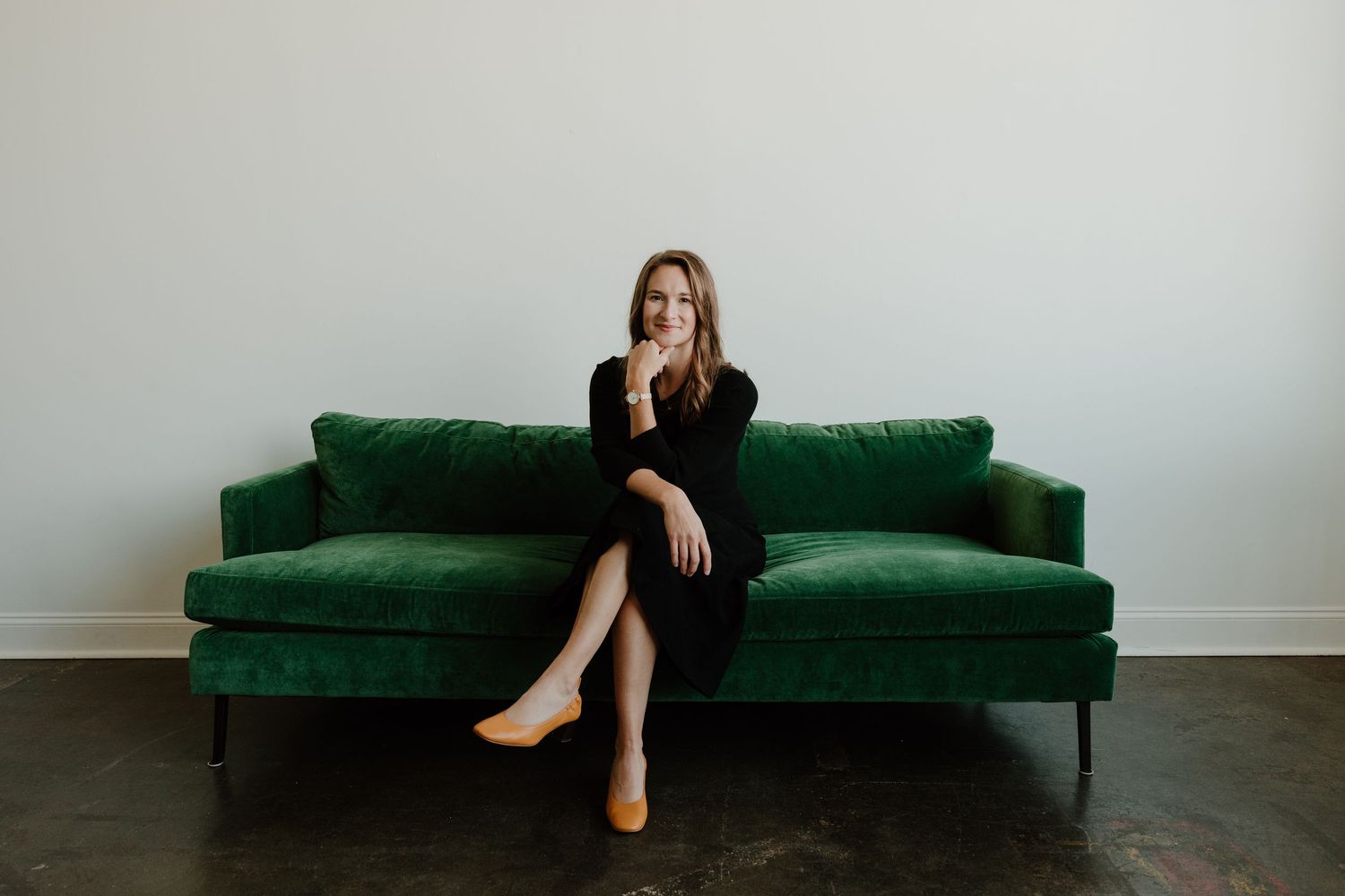 A person in a black dress sits elegantly on a luxurious emerald green velvet sofa against a white wall.