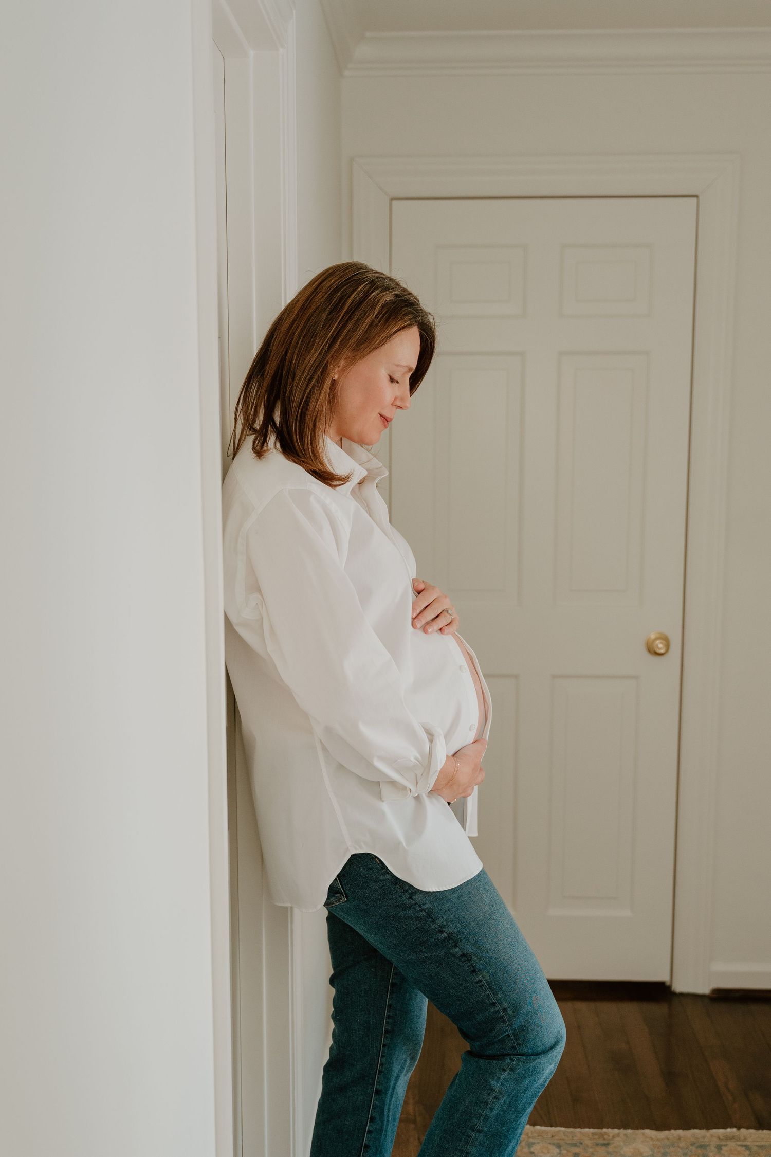 Woman in white button-down shirt and teal pants posing playfully against a white wall.