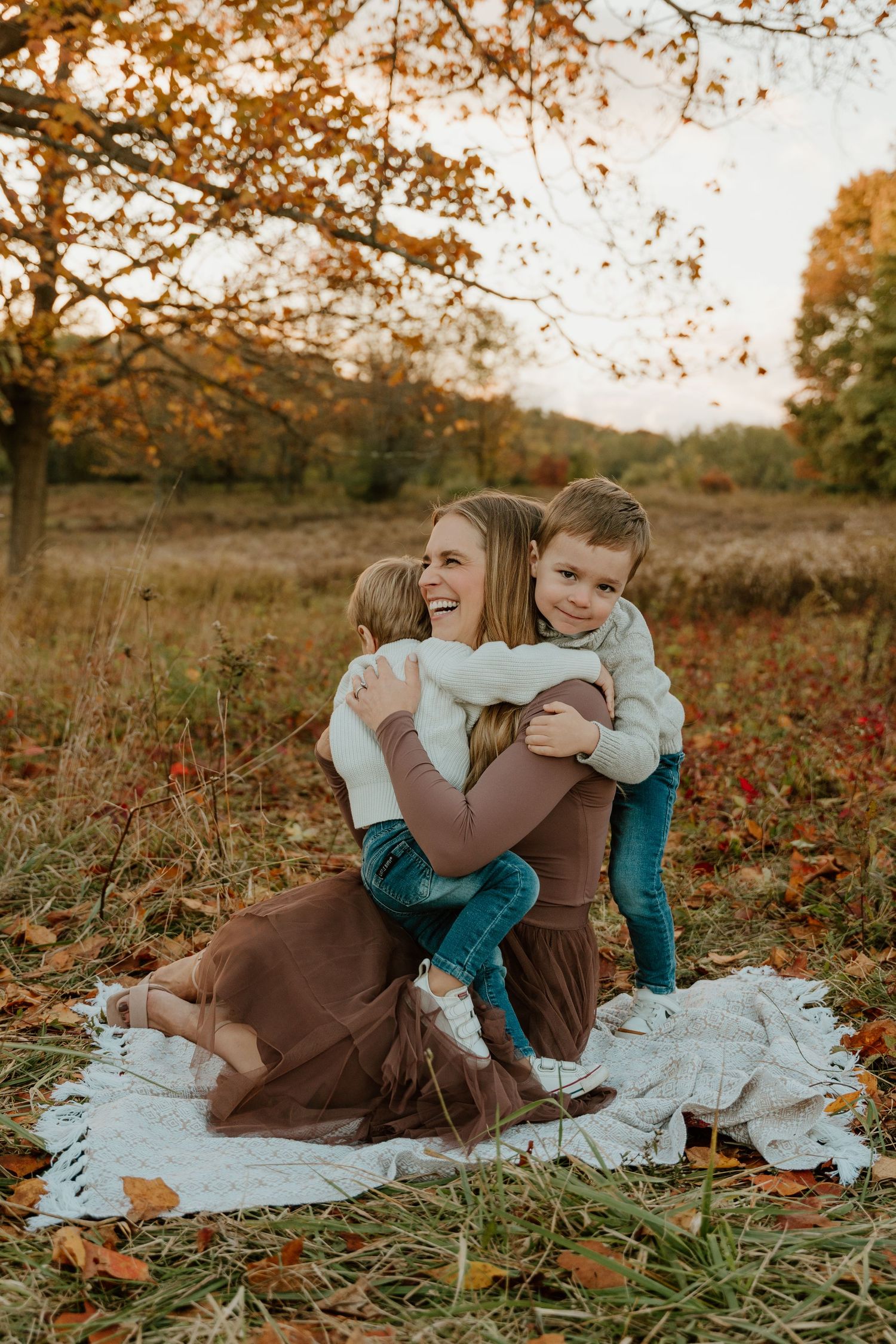 A family enjoys a warm autumn day sitting on a blanket in a field surrounded by golden fall foliage at sunset.