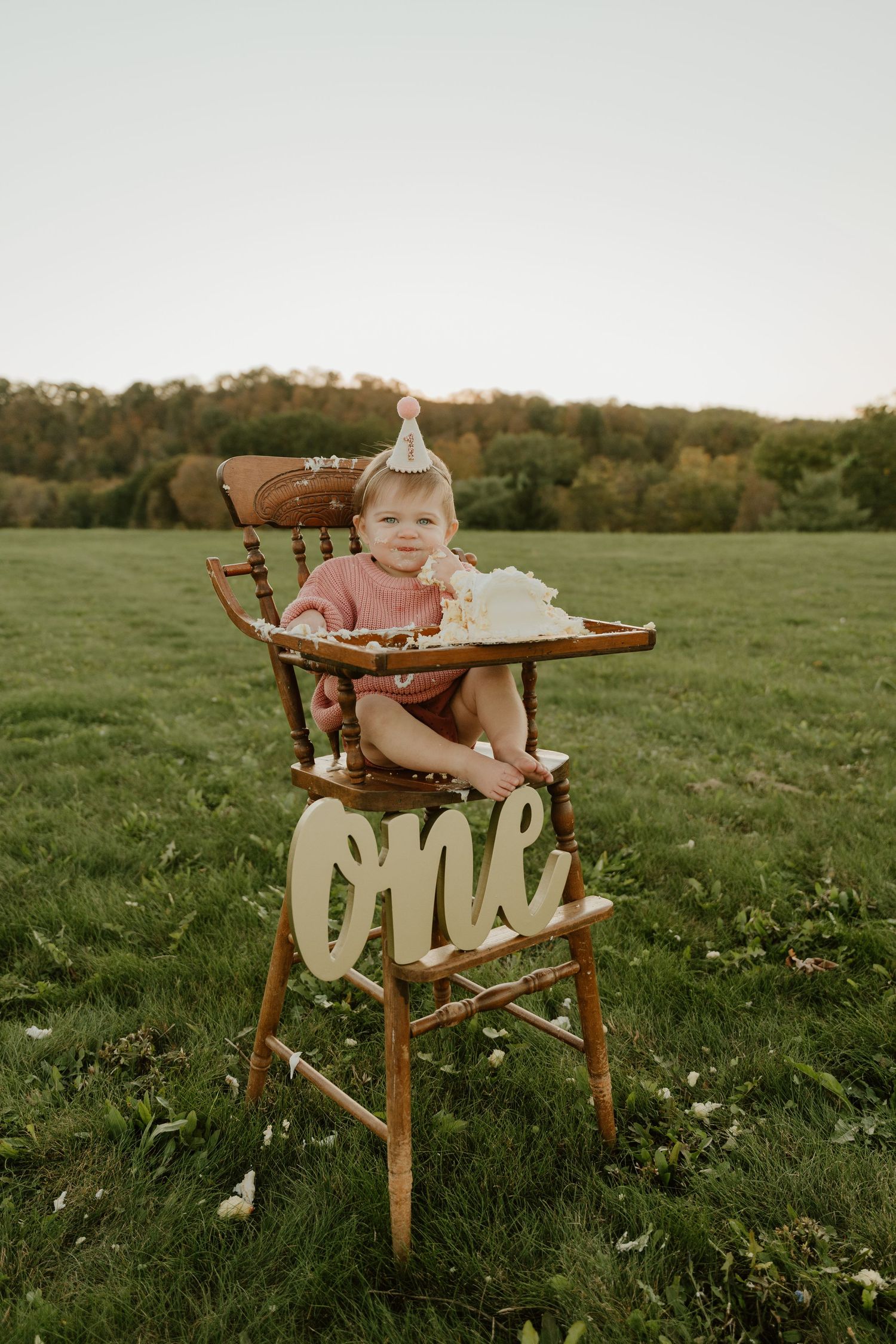 Vintage wooden high chair with 'ONE' decoration sits in a grassy field during golden hour.
