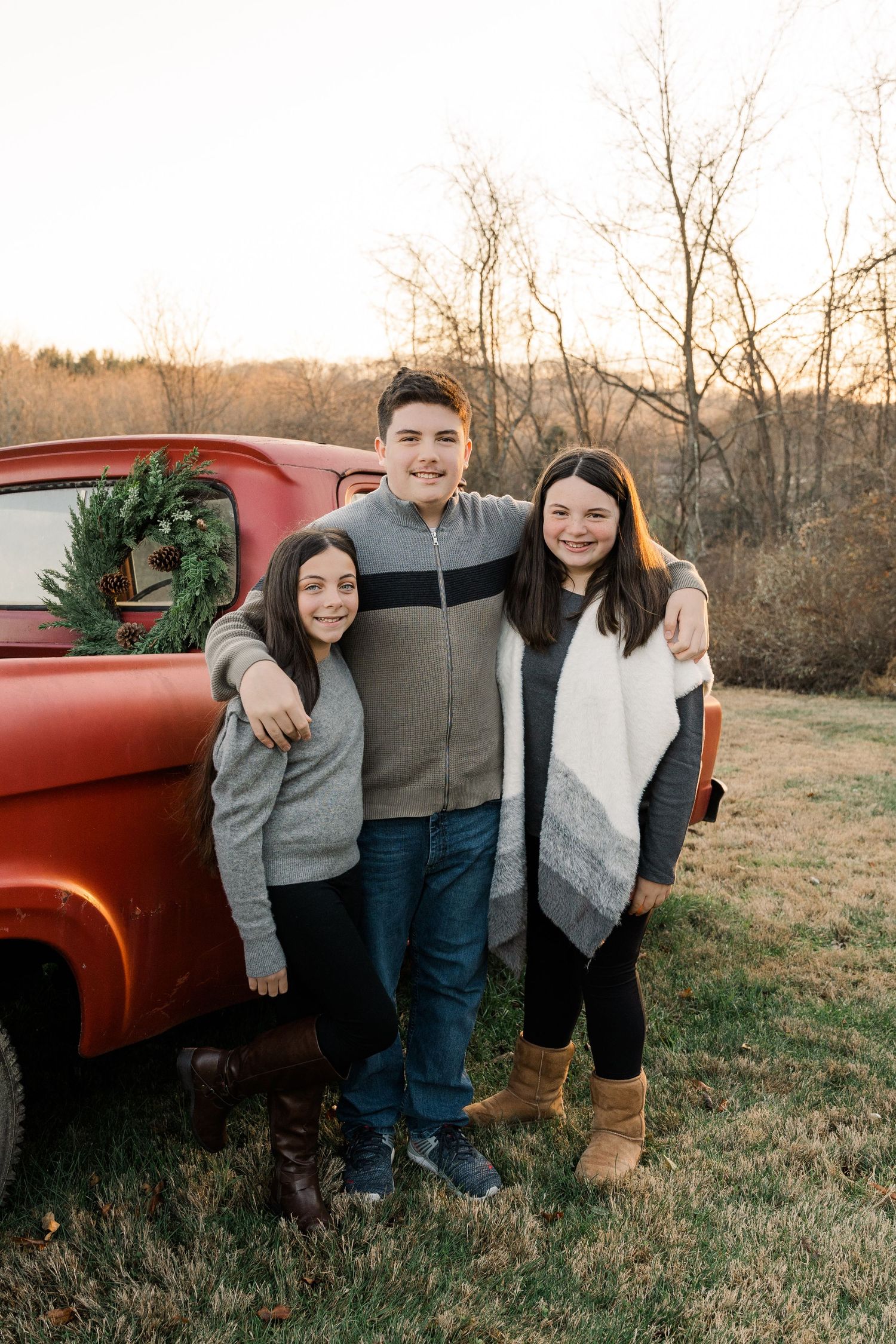 Three siblings pose together by vintage red truck with holiday wreath.