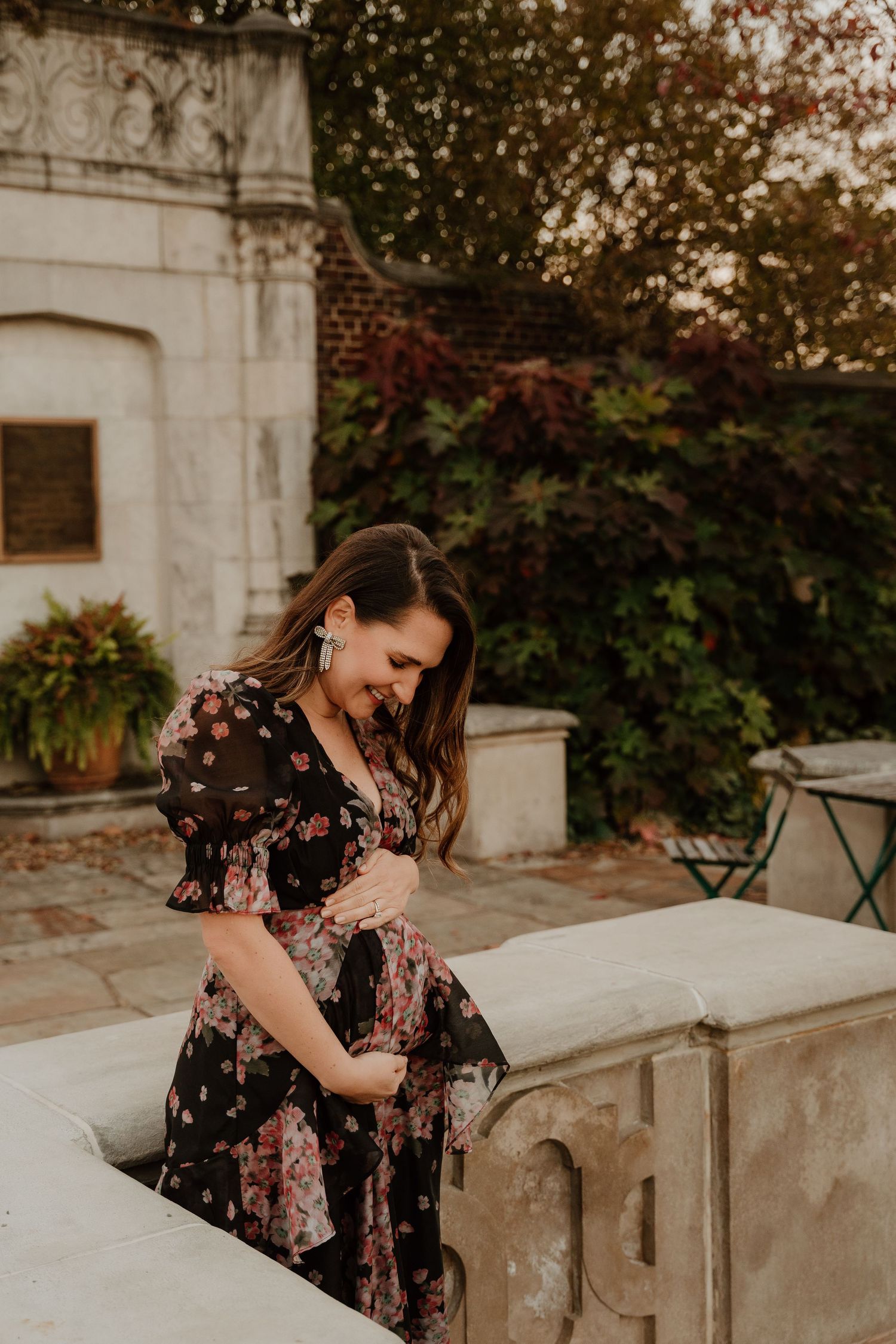 Woman in floral dress smiling while sitting on stone wall in garden setting.
