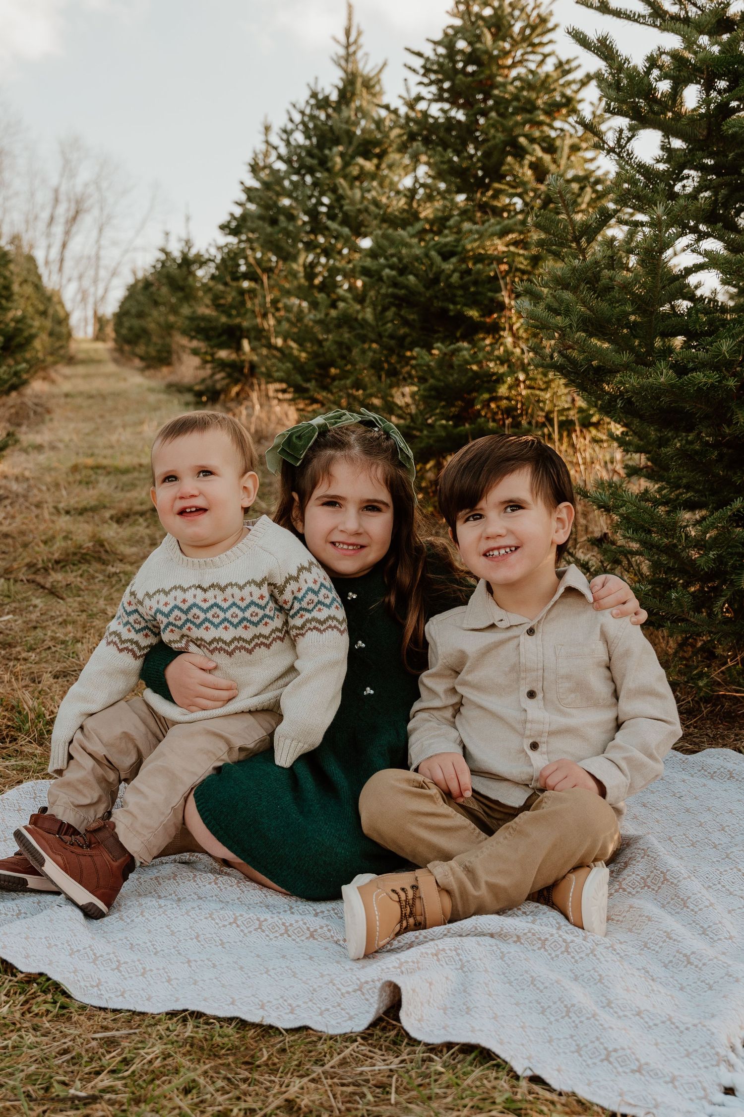 Three children sitting together on a white blanket outdoors among evergreen trees at Candletree Farm at sunset.