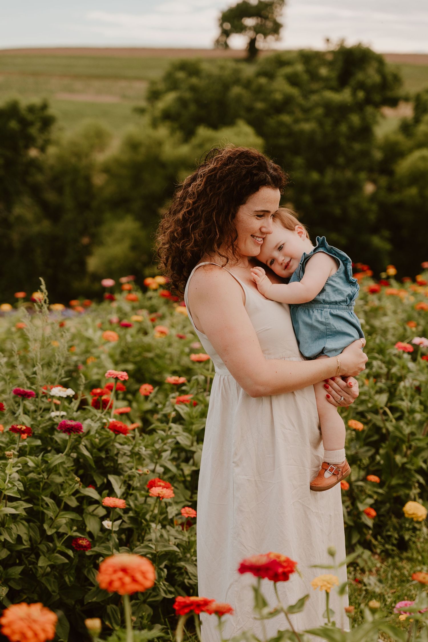 A mother in a white dress holds her smiling toddler in a vibrant zinnia flower field outdoors.