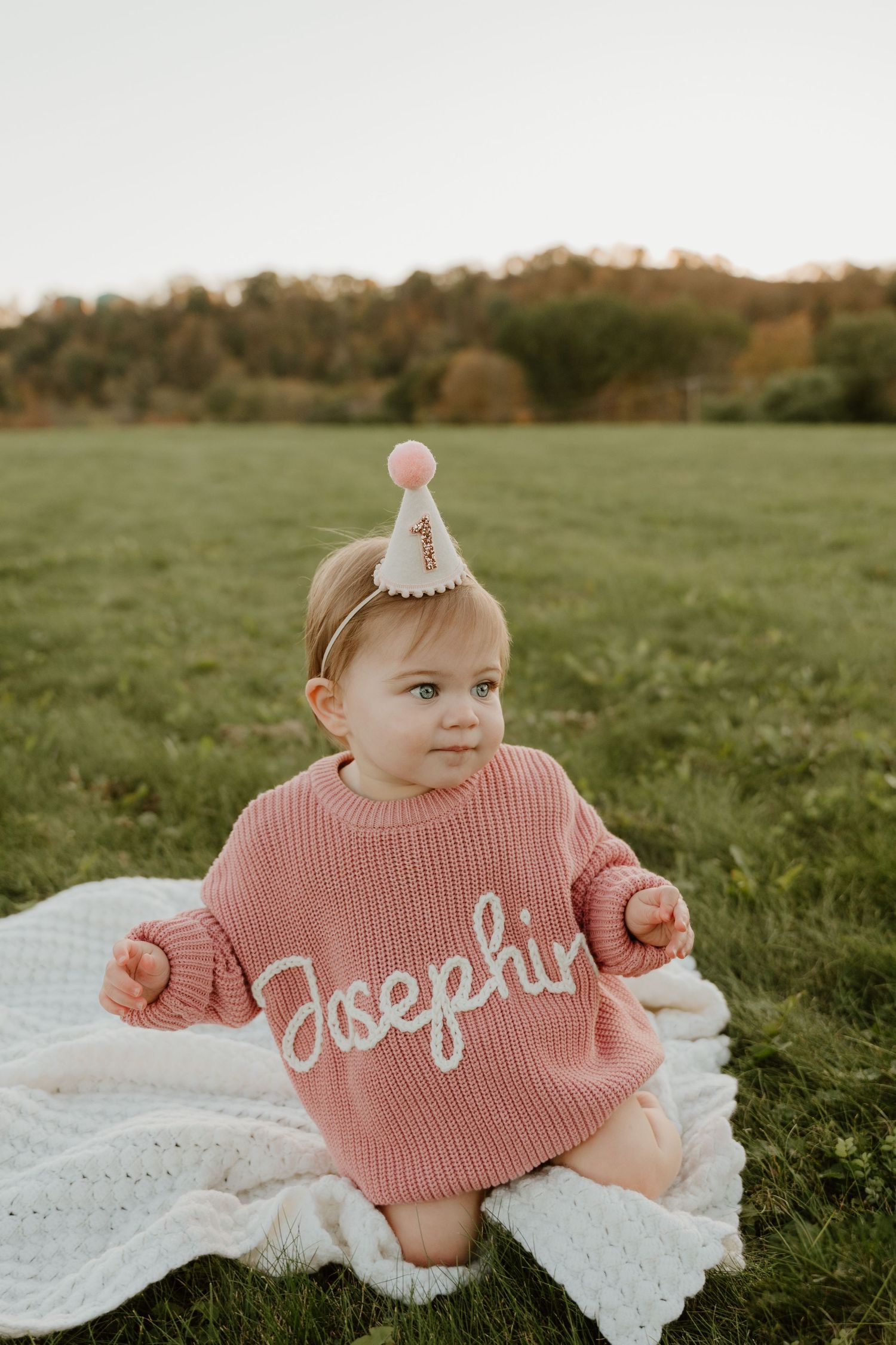 A baby in a pink knit sweater sits on a white blanket in a grassy field during golden hour.