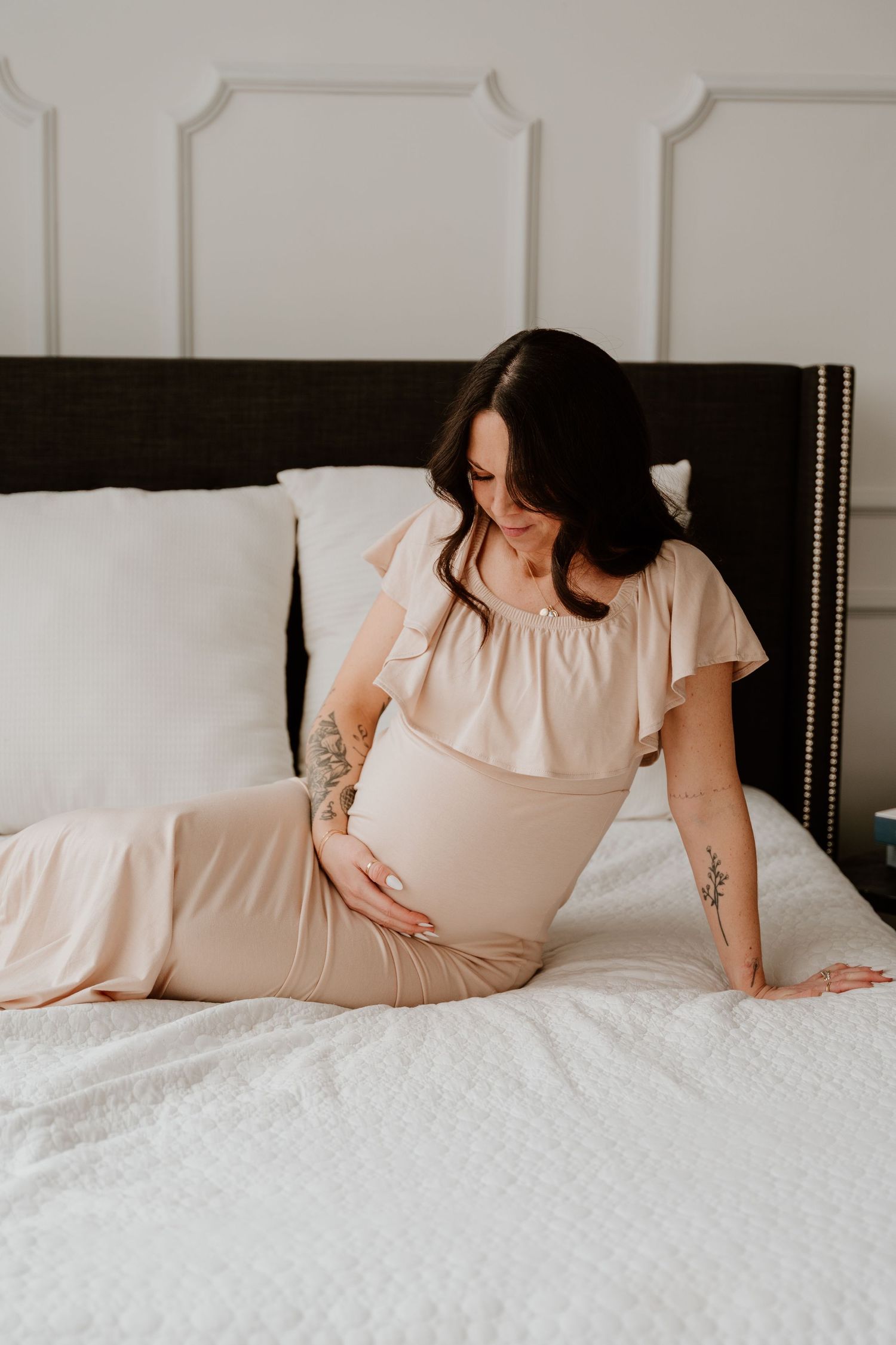 Pregnant woman in beige dress sitting on white bedding looking down at her baby bump in a cozy bedroom setting.