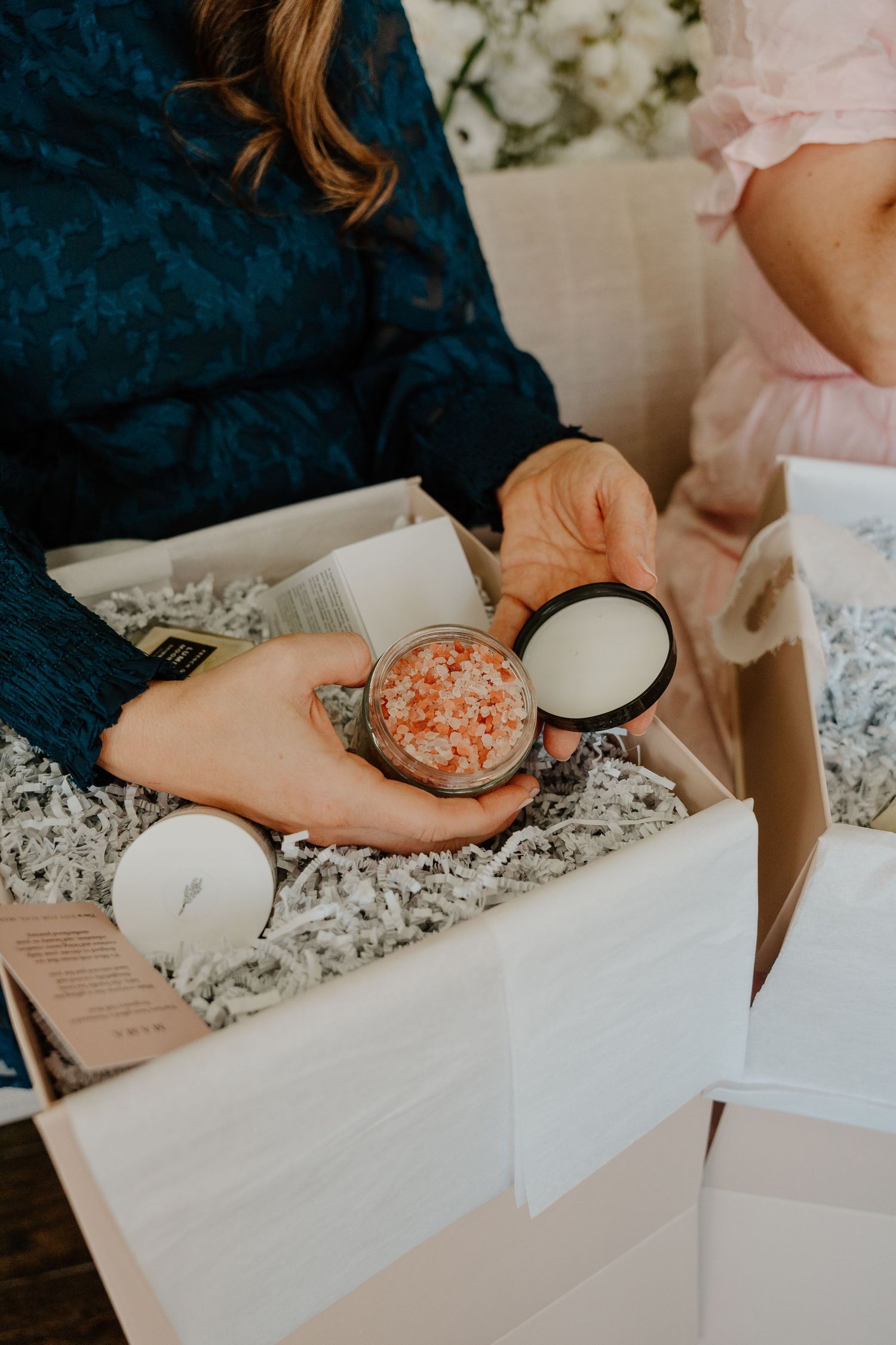 Hands holding a pink bath bomb while writing on paper with other bath products and packaging materials on a white surface.